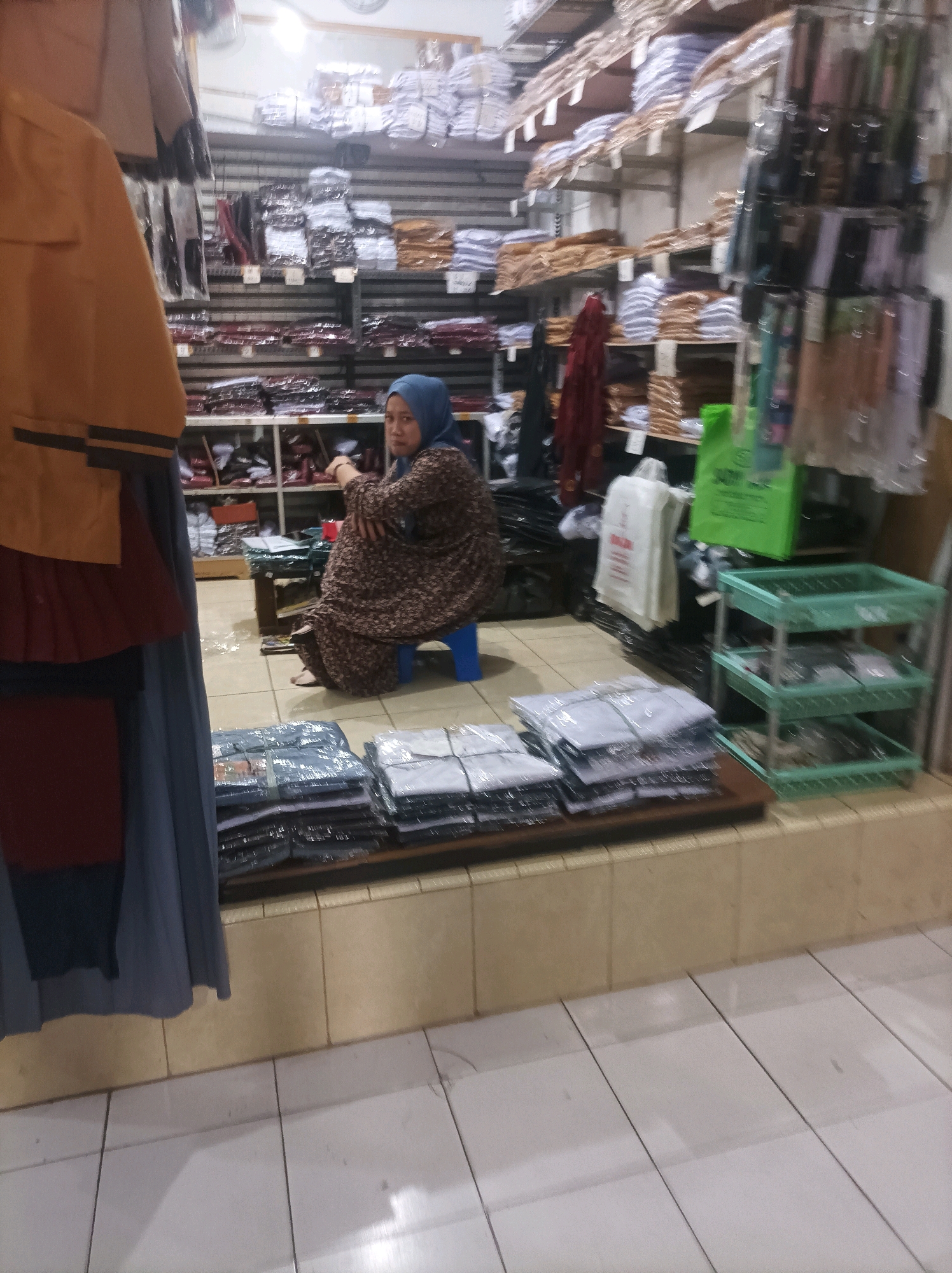 A person wearing a blue hijab and a patterned brown dress sits on a blue plastic stool in a store, surrounded by shelves of folded clothing. To the left, a section of yellow, red, and blue fabric hangs, likely part of a garment. In the foreground, stacks of folded, packaged clothing are arranged on shelves. A green plastic rack with multiple shelves sits to the right of the packaged clothing. The floor is tiled with white and beige squares. The store appears to be a retail establishment selling apparel in Sumuragung, Indonesia.