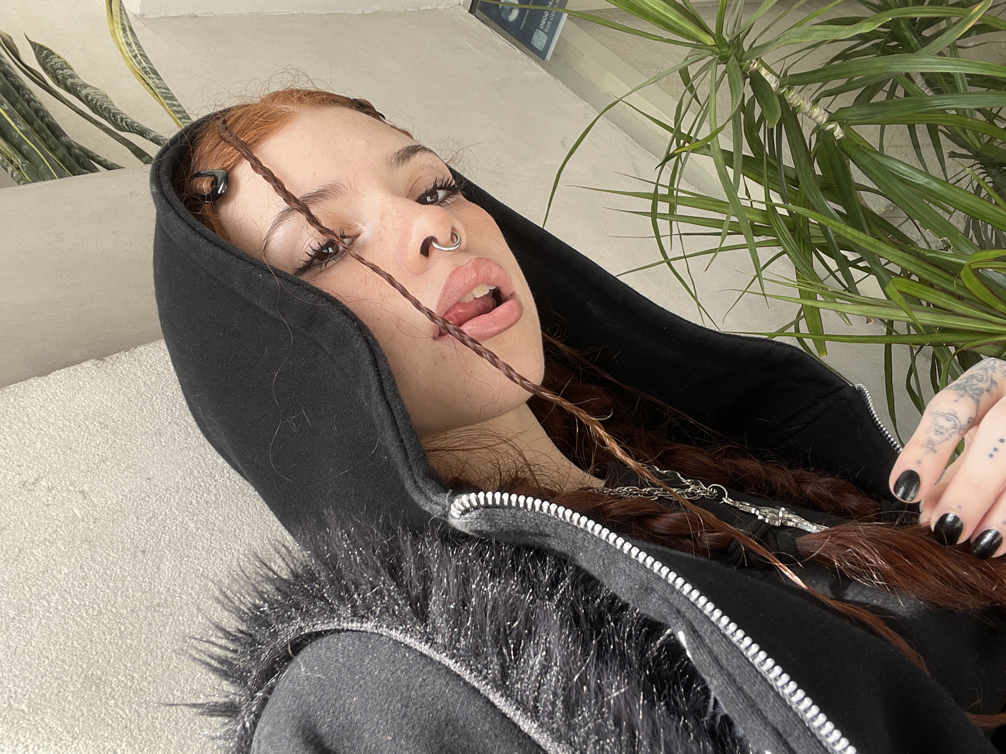 A selfie perspective shows a young woman indoors. She is wearing a black hooded jacket with what appears to be fur trim. Her hair is reddish-brown, styled in braids with dark hair clips. She has facial piercings on her nose and is wearing dark nail polish. Her skin tone appears fair. Parts of two plants are visible in the background, a snake plant to the left and a dracaena to the right. The background wall is white, possibly in an apartment setting. There is nothing in the image to associate the image with Buenos Aires, Argentina.