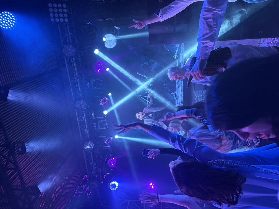 The image captures an indoor live performance event in Редант, Russia. A male performer with dark hair, wearing a grey jacket over a dark shirt, stands center-stage, mid-action with arms spread and mouth open, indicative of singing or engaging the audience. He is positioned in front of a large screen or projection displaying a blue-toned, possibly lunar, landscape with prominent white stylized Cyrillic letters "А А". Above the stage, a truss system supports numerous blue and purple LED lights and spotlights, casting visible beams through a hazy atmosphere. The audience occupies the foreground and midground, largely obscured but demonstrating active participation: multiple hands are raised, some individuals are pointing towards the stage, and one person holds a smartphone upward, likely recording. A grey-haired man in a light blue shirt is visible in the mid-right, looking towards the stage. A large dark speaker cabinet is situated on the right side of the stage. The venue's ceiling exhibits a corrugated metal structure. Confetti or small reflective debris is scattered on the floor in the immediate foreground.