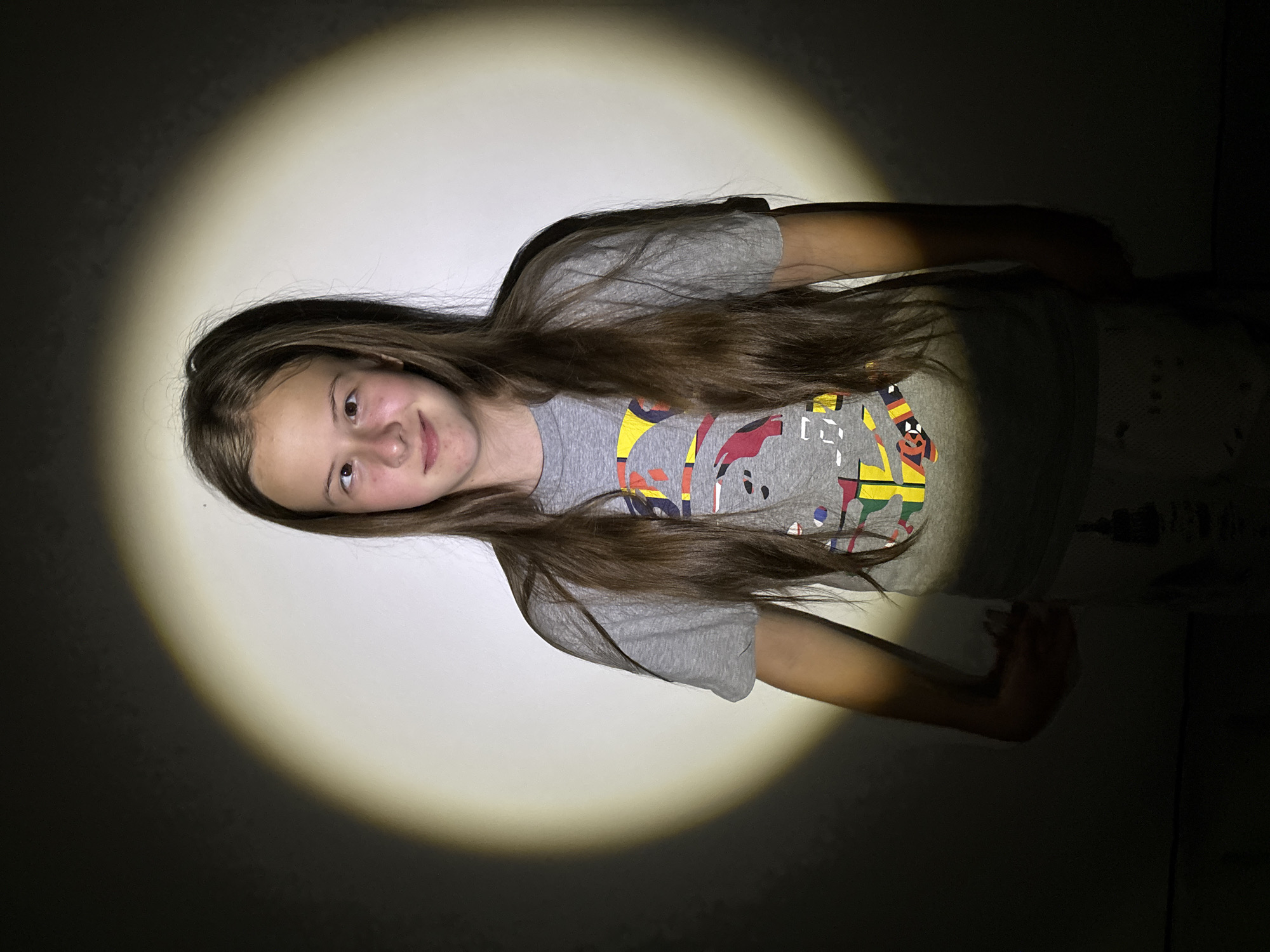 A young girl, appearing to be in her early teens, is featured in a half-body portrait. She has long, dark brown hair that flows over her shoulders and chest. She is wearing a grey t-shirt with a colorful, abstract graphic design on the front, which includes various shapes, patterns, and stylized elements resembling letters or cartoon figures in shades of yellow, red, blue, and black.
The girl is illuminated by a prominent, circular spotlight that casts a bright glow on her and the plain wall behind her, creating a strong contrast with the dark surroundings. She looks directly at the viewer with a subtle, pleasant smile. The focused lighting suggests the photograph was taken indoors in a low-light environment, likely at night. The location context for this image is Chaikovsky, Russia.