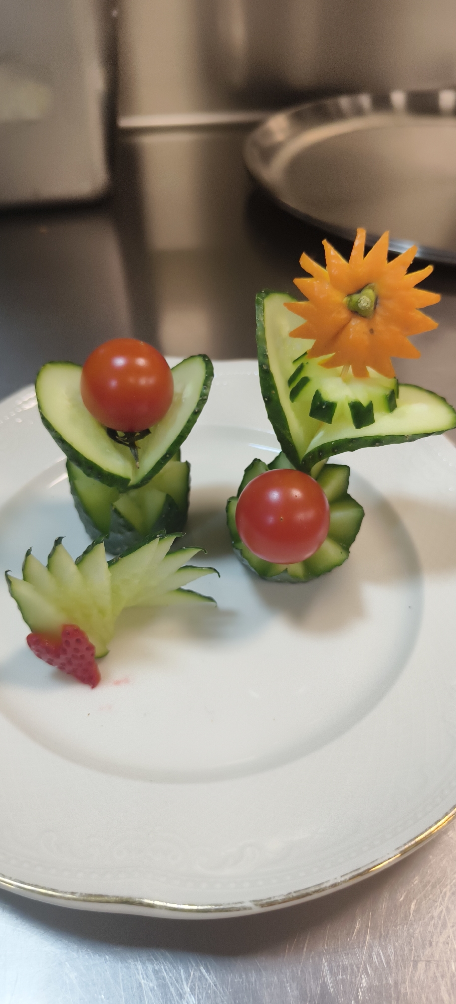 This media file features a close-up shot of elaborately carved vegetables presented on a white plate. The main subjects are three decorative food arrangements, primarily made from fresh cucumber, cherry tomatoes, and carrot.
Two of the arrangements consist of green cucumber slices intricately carved and folded to create a base, each cradling a single bright red cherry tomato at its center. The arrangement on the right is further adorned with a vibrant orange carrot, expertly carved into a spiky, star-like flower that sits atop the cucumber structure. This particular cucumber also features small, square cutouts, adding to its decorative appeal.
A third, smaller decorative element is positioned on the lower left of the plate. It showcases a fanned-out cucumber carving, resembling a delicate leaf or a small frond, with a small piece of red strawberry cut into a heart shape placed at its base.
The white plate has a subtle, embossed pattern along its rim and a thin gold border, slightly worn in places. The background is blurred but appears to be a metallic surface, suggestive of a kitchen counter or serving area, reflecting ambient light. The overall scene highlights the art of food garnishing and presentation, showcasing skilled culinary craftsmanship.
No people are visible in the image, and there is no visible text. The image originates from Yerevan, Armenia, although no specific location cues are present within the frame itself. The lighting appears to be indoor and well-distributed, illuminating the food preparations clearly.