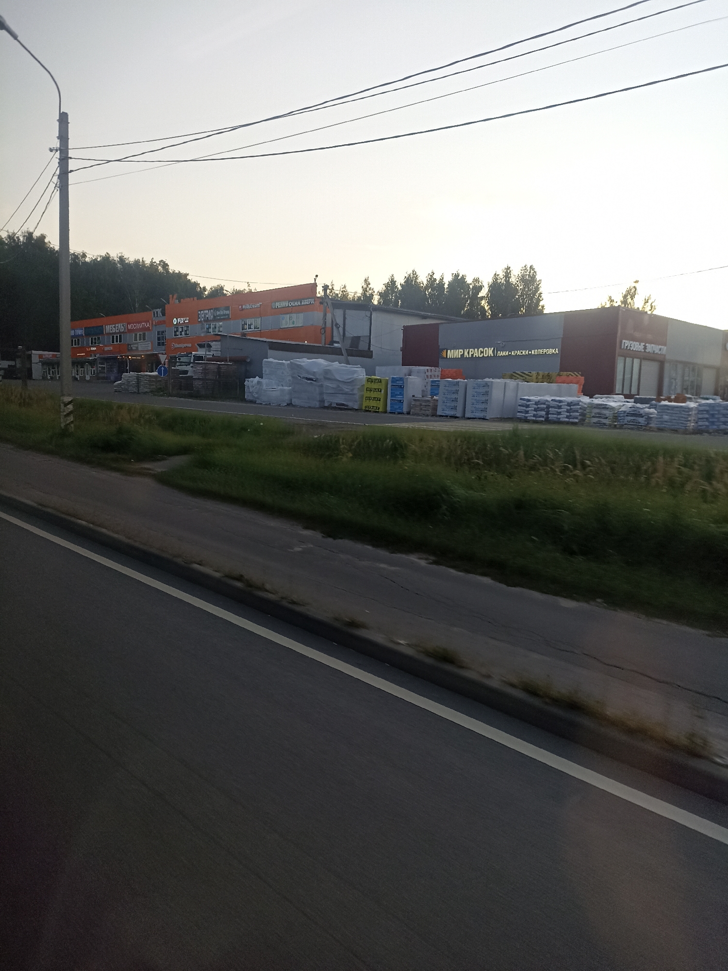 The image depicts a commercial building with multiple businesses on the left side of the frame, likely situated along a road in Sandarovo, Russia. The building features an orange facade with various signage, including "МЕБЕЛЬ МОСПЛИТКА" (Furniture Mosplitka), "ЭЛЕКТРИКА" (Electrics), and "FIX PRICE" (a discount retail chain). The right side of the building has a darker facade with signage that appears to read "МИР КРАСОК" (World of Paints) followed by "ЛАКИ КРАСКИ КОЛЕРОВКА" (Varnishes, Paints, Coloring), and further to the right, "ГРУЗОВЫЕ ЗАПЧАСТИ" (Truck Parts). In front of the building, there are stacks of white and grey materials, possibly construction or packaging supplies. The scene is viewed from a moving vehicle, indicated by the blurred asphalt road in the foreground and the sense of motion. The sky is overcast, suggesting it is either early morning or late evening. Trees line the background behind the buildings. There are no people clearly visible in the image.