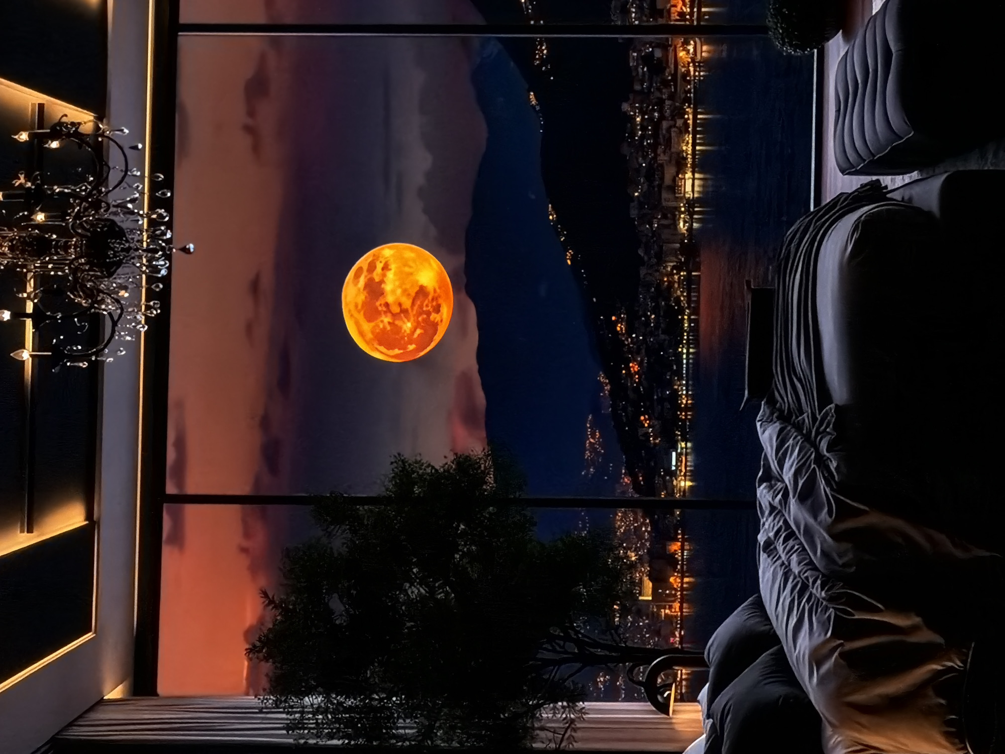 This image presents a dramatic nocturnal scene viewed from a modern, luxurious interior through large windows. The overall impression is one of opulence and a grand vista.
Inside the room, visible elements suggest a sophisticated living space. On the far left, a grand, ornate chandelier with numerous light bulbs and delicate crystal drops hangs, contributing to the room's elegant ambiance. Below it, a contemporary wall panel with warm, illuminated strip lighting runs vertically, casting a soft glow. To the right, dark, plush furniture, possibly a sofa or armchair, is partially visible, draped with soft fabric, indicating comfort. A dark, leafy plant or small tree stands silhouetted near the window in the lower center.
Beyond the window, a breathtaking night landscape unfolds. Dominating the center-left is a colossal, intensely glowing orange full moon, detailed with a textured surface resembling craters, giving it a powerful, almost fiery appearance. The sky behind the moon transitions from a warm orange-red hue on the far left, suggesting a late twilight or afterglow, to a deep, dark indigo blue on the right, indicating night. Silhouetted mountains or hills rise majestically under the moonlight. Below these landforms, a vast body of water, likely a lake or sea, stretches across the view, reflecting the faint light from the sky and the distant urban glow. Along the far shore and extending into the hills on the right, a sprawling cityscape or town twinkles with countless small, golden lights, creating a vibrant and extensive night skyline.
The scene captures a peaceful yet dramatic moment at night or during deep twilight. No people are visible, and there is no visible text. The atmosphere is one of serene luxury and a slightly fantastical grandeur, with the oversized, vibrant moon being a focal point. The location context is Sofiivska Borshchahivka, Ukraine.
