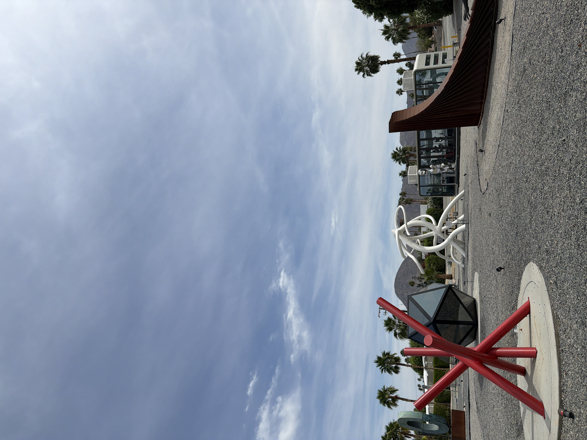 The image depicts a public space in Palm Desert, California, featuring several modern art installations. A prominent sculpture in the foreground consists of red cylindrical rods intersecting in an abstract design, set atop a circular concrete base. Behind it, there's a dodecahedron-shaped structure with reflective panels. Further back, a white, curvilinear sculpture stands out, followed by an art installation featuring a series of curved, rusted metal plates. Palm trees are interspersed throughout the scene. The ground is covered in small, dark pebbles. In the background, there is a partially visible white building with visible reflective windows and palm trees. A clear sky with wispy clouds is present.