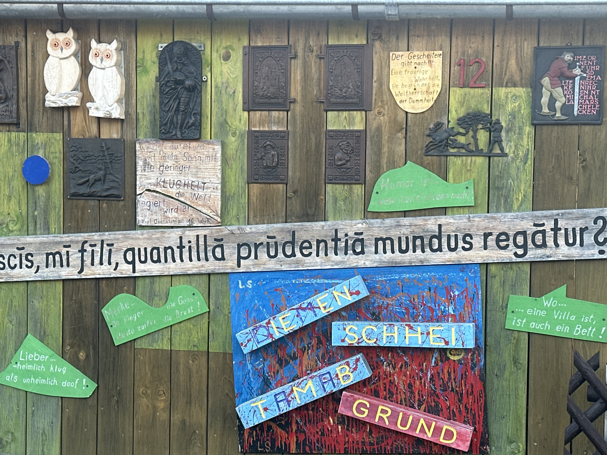 The image shows a rustic wooden wall adorned with various decorative elements and inscriptions, located in Günzburg, Germany. The wall is constructed from vertical wooden planks with a weathered, green and brown patina.
Prominently displayed are two carved wooden owls, facing left and right respectively, mounted at the top left. To their right is a dark, rectangular plaque featuring a carved figure of a bearded man holding a sword and a book. Further to the right, a sign with the number "12" in red is visible. Below the owls and to the left, a dark plaque depicts a hunting scene with a man aiming a rifle at a deer. Another dark plaque, to the right of the hunting scene, features four smaller framed reliefs, two at the top and two at the bottom, each with a figure.
Several wooden signs with text are also present. One, a light-colored wooden plank with dark lettering, reads: "Wer den Schein mit geringer KLUGHEIT die Welt regiert wird 21." Below this are the initials "P.REL. Oxenstierna. 16. Jahrh." To the right of this sign is a green, fish-shaped sign with the word "Humor" in a lighter shade, followed by "ist...wenn man's a-r-d-e-n mach!"
A large, horizontal wooden sign spans the lower portion of the image, bearing the Latin inscription "scis, mi fili, quantilla prudentiā mundus regātur?".
Below the Latin inscription, a vibrant, abstract painting with splashes of blue, red, and yellow serves as a backdrop for several wooden lettered signs. These signs, arranged diagonally, spell out "DIETEN SCHHEIT", "TAMAB", and "GRUND" in yellow letters against a blue painted background with red and black paint splatters.
To the left of the abstract painting, a green, curved sign reads: "Lieber ... heimlich klug als unheimlich doof!". To the right of the painting, another green, curved sign states: "Merke.. je jünger desto zaffer die Brust!". And further to the right, a green, pointed sign reads: "Wo...eine Villa ist, ist auch ein Bett!".
The environment suggests an outdoor setting, likely a garden or a decorative outdoor wall, given the natural materials and the decorative nature of the objects. The style of the decorations suggests a whimsical and perhaps historical or literary theme.
