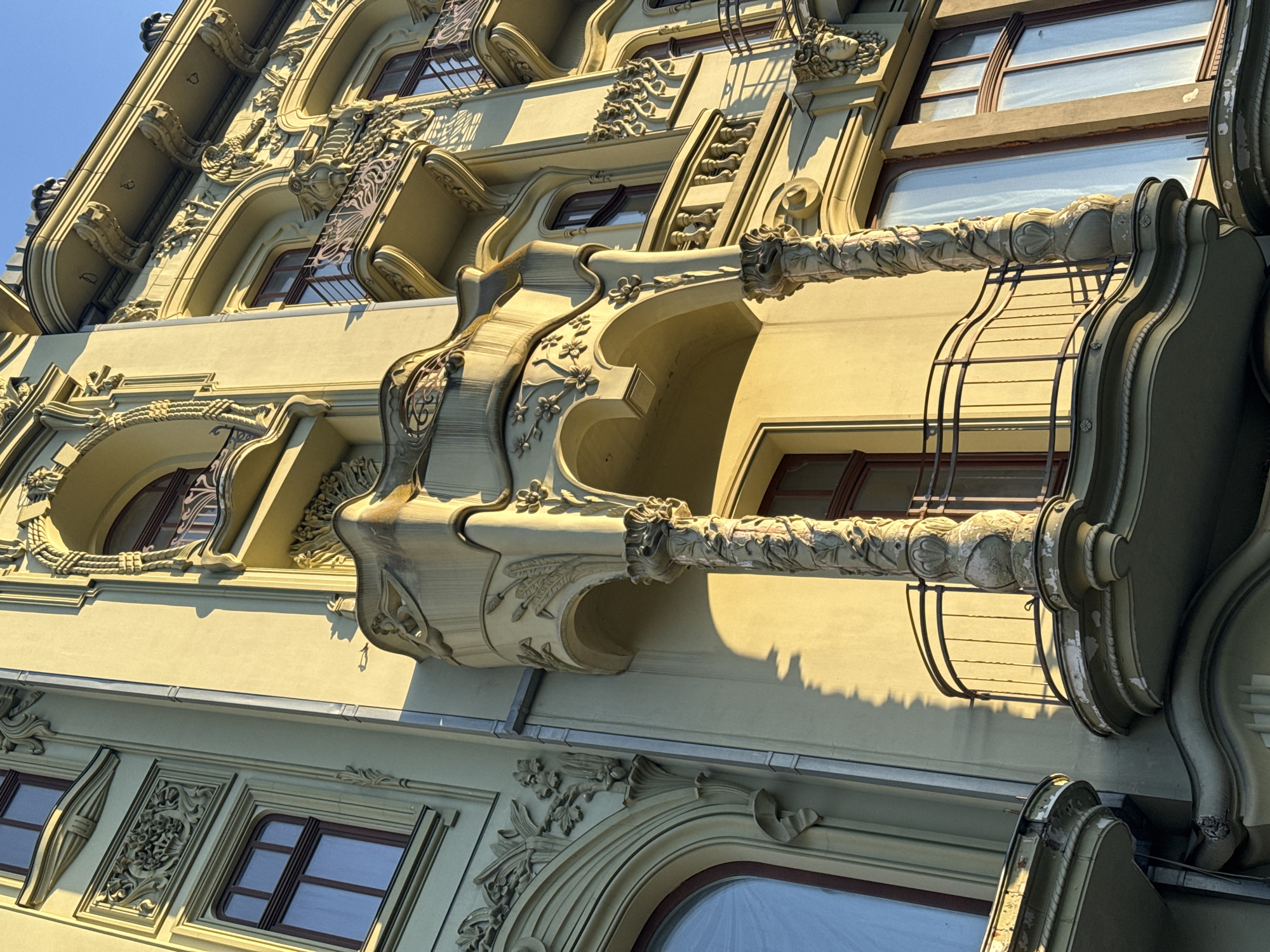 The image shows a close-up, low-angle view of the facade of the elegant Odessa National Academic Theater of Opera and Ballet, located in Odesa, Ukraine. The sun is casting long shadows across the building's ornate, art nouveau architectural details, highlighting the intricate carvings, balconies, and windows. The building's exterior is painted in a pale yellow or lime green hue. The sky above is a clear, bright blue. The focus is on the architectural features, with no people or activity visible. The detailed ornamentation, including floral motifs, curved lines, and sculptural elements, is a prominent characteristic of the building's design.