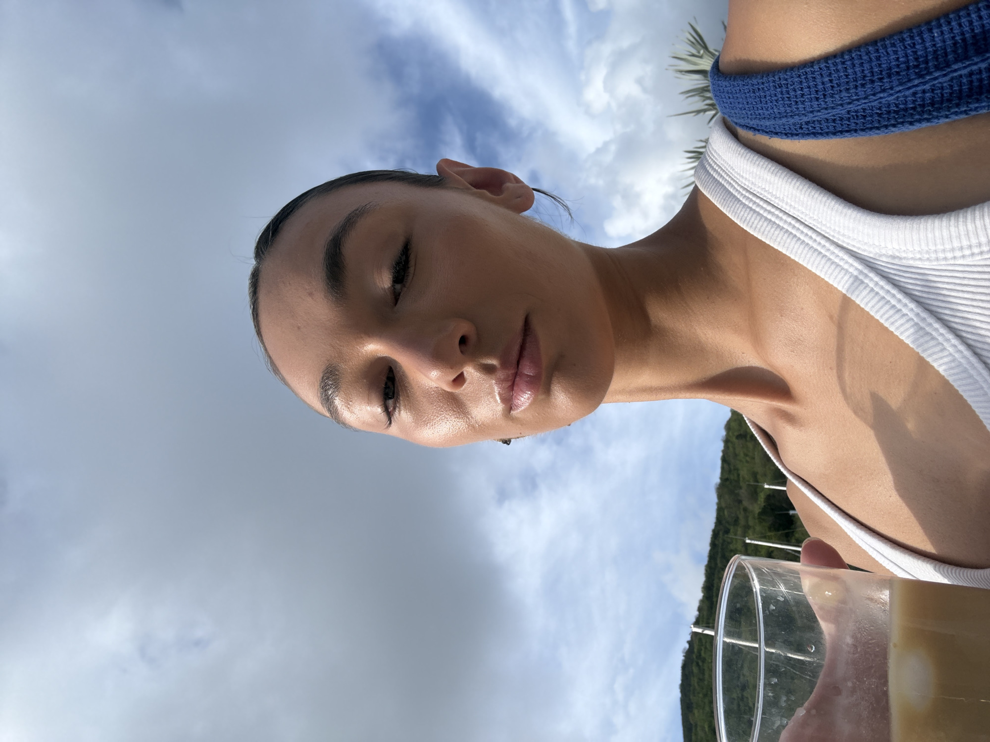 A young woman is captured in a low-angle close-up shot, looking downwards and slightly to her right. Her face is well-lit by natural light, highlighting a serene expression, dark, defined eyebrows, and full, glossy lips. Her dark hair is pulled back, revealing her ears. She wears a white ribbed tank top, and a textured blue strap, possibly from swimwear, is visible over her right shoulder.
She holds a clear glass containing a light yellow or orange beverage with ice cubes in her right hand, visible in the lower right corner. The background is dominated by a bright sky with scattered white and grey clouds. On the lower right side, a lush green, tree-covered hillside or mountain range can be seen, hinting at a natural, possibly tropical environment. Faint structures, resembling utility lines or parts of a bridge, are discernible against the green landscape.
The image suggests a daytime setting with clear but partly cloudy weather. The overall ambiance is relaxed and bright, consistent with a vacation or leisure activity in a scenic location like Saint-Martin, France. There is no visible text in the image.