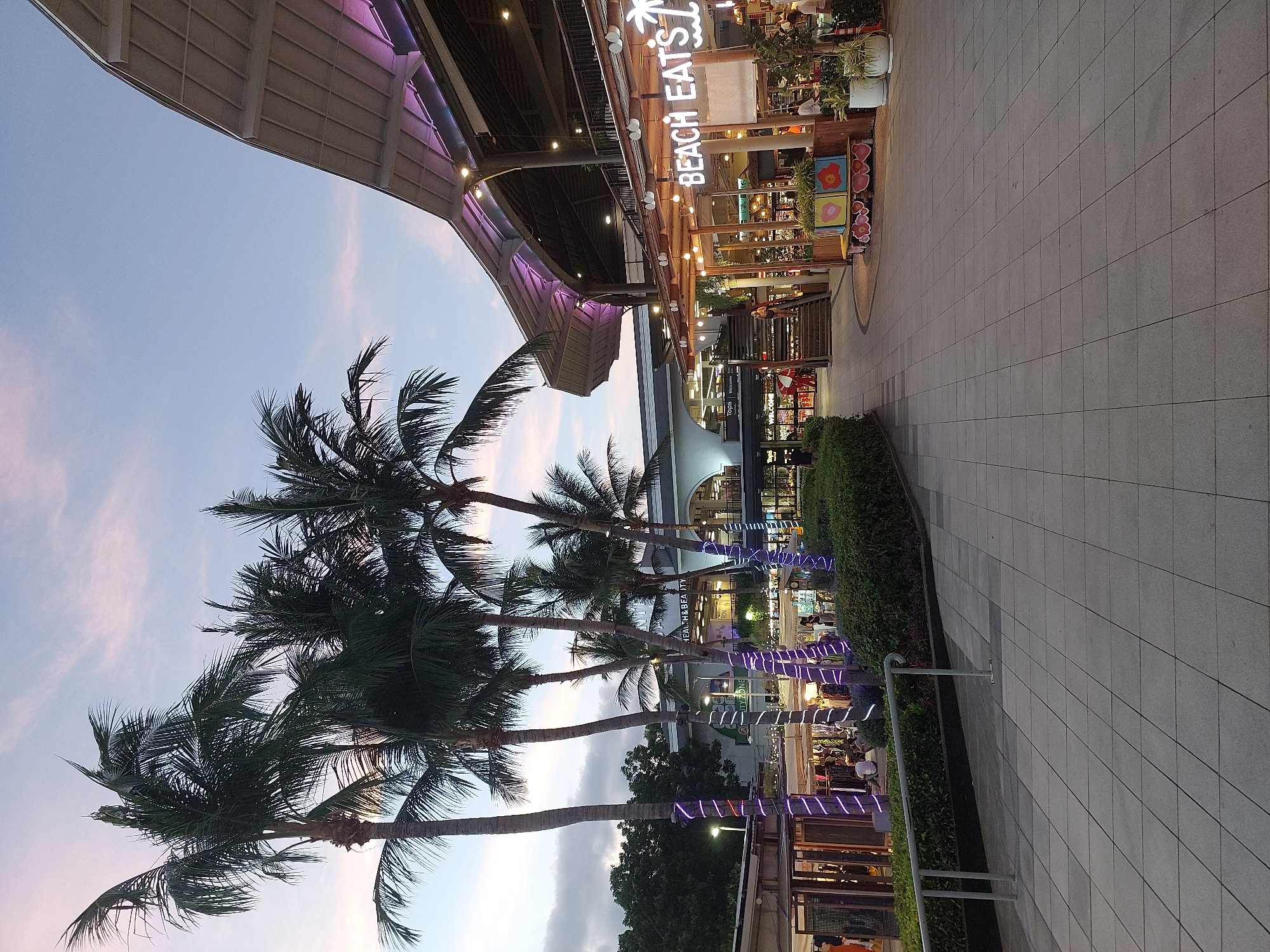 The image depicts an outdoor walkway at dusk, with a clear sky transitioning into evening. Several palm trees stand tall, their fronds casting shadows across the tiled pavement. The walkway is lined with shops and restaurants, their facades illuminated by various lights. Some of these establishments feature outdoor seating areas where people can be seen dining and socializing.
A prominent sign reads "BEACH EATS" in neon lettering, indicating a food-related establishment. Decorative lights, some appearing as purple and white stripes wrapped around poles, add a festive ambiance to the scene. The architecture suggests a modern commercial area, possibly a beachfront promenade or a shopping complex designed to evoke a tropical atmosphere. The overall mood is relaxed and inviting, suggesting a pleasant evening out. The time of day is indicated by the soft, diffused light and the illuminated signs and shopfronts, suggesting the transition from sunset to evening. The weather appears to be clear, with a few wispy clouds visible in the sky. While no specific event is clearly taking place, the scene conveys a sense of everyday evening activity in a popular public space.