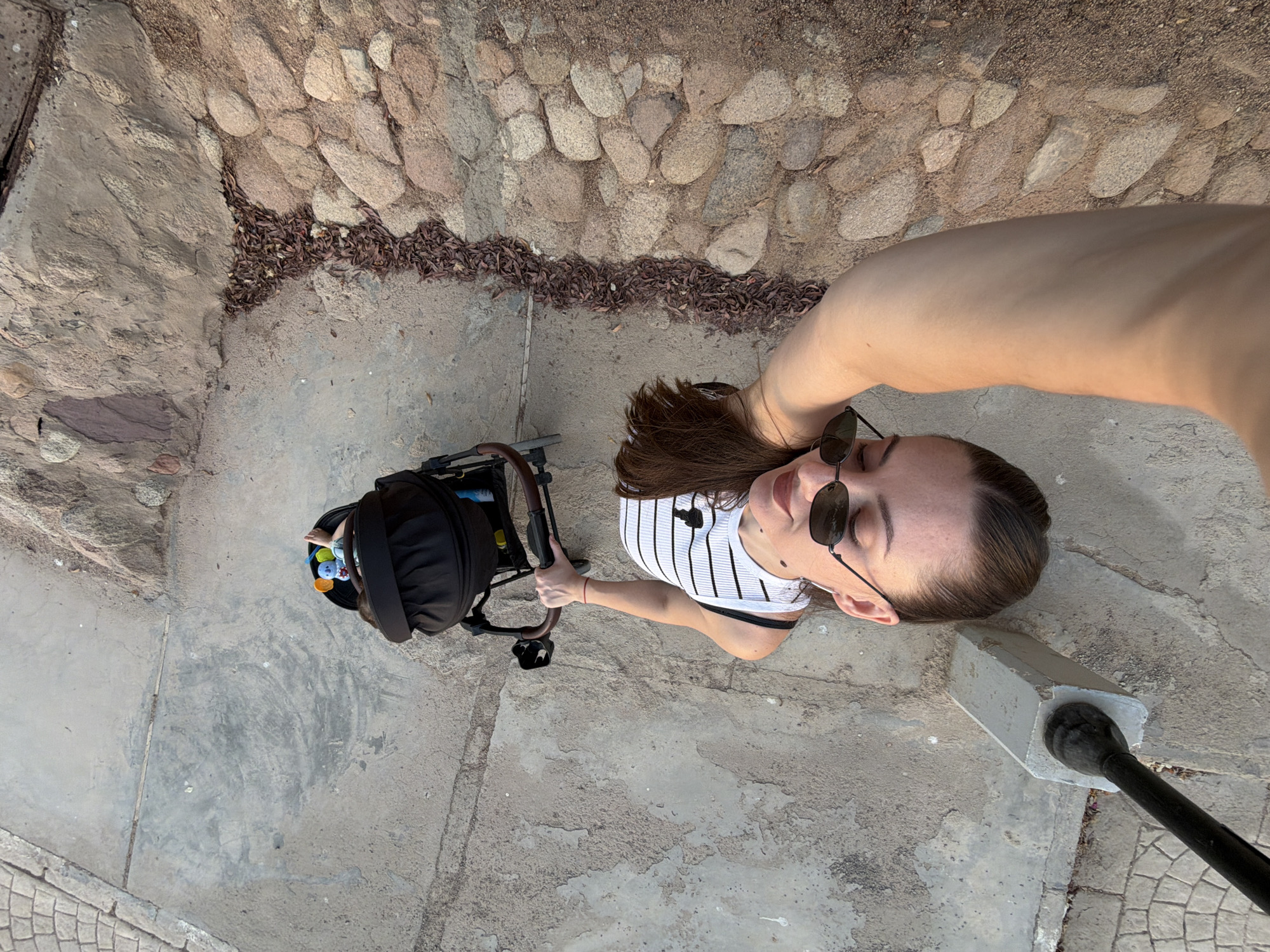 A top-down, high-angle selfie captures a young woman pushing a black baby stroller. She is wearing a white and black striped tank top and dark sunglasses. Her hair is dark brown and pulled back. The stroller contains a baby, whose feet are visible, along with a colorful toy attached to the stroller's canopy. The woman's right arm extends upwards, holding the stroller's handle, and her left arm appears to be holding the camera, which is out of frame. The ground is a mix of concrete paving stones and natural, rounded stones. A pile of dry leaves is visible between the two ground surfaces. In the foreground, a section of a concrete structure with a black metal pole protruding from it is partially visible. The image appears to be taken outdoors, possibly on a promenade or walkway in Sharm El Sheikh, Egypt, given the distinctive paving and rocky terrain.