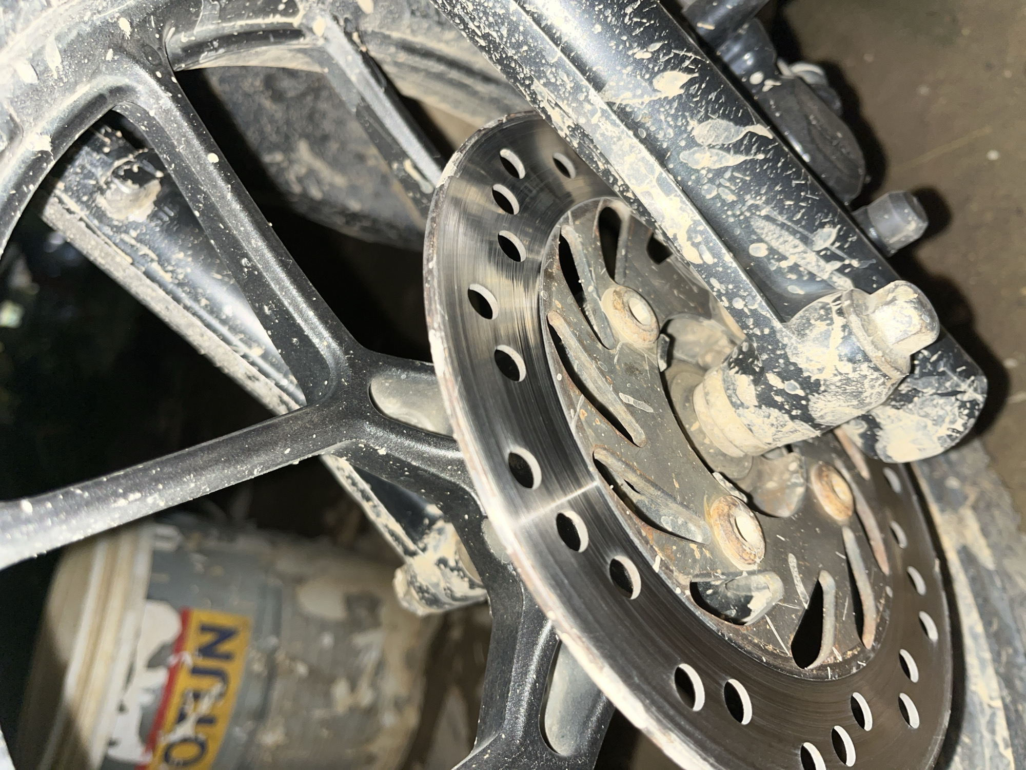 The image shows a close-up of a motorcycle's rear brake disc and wheel, heavily splattered with mud. The disc itself is a circular metal plate with several holes drilled into it, showing signs of wear and some rust. The wheel is black and appears to be made of a cast alloy, with multiple spokes. The surrounding frame and suspension components are also covered in mud, suggesting the motorcycle has been used in wet, off-road conditions. In the background, a partially visible bucket with a faded yellow and red label, possibly containing paint or another substance, is also present. The environment appears to be an outdoor or semi-outdoor space, with the ground covered in dirt and debris. The lighting is dim, highlighting the texture of the mud and metal surfaces. There are no people or animals visible in the frame.