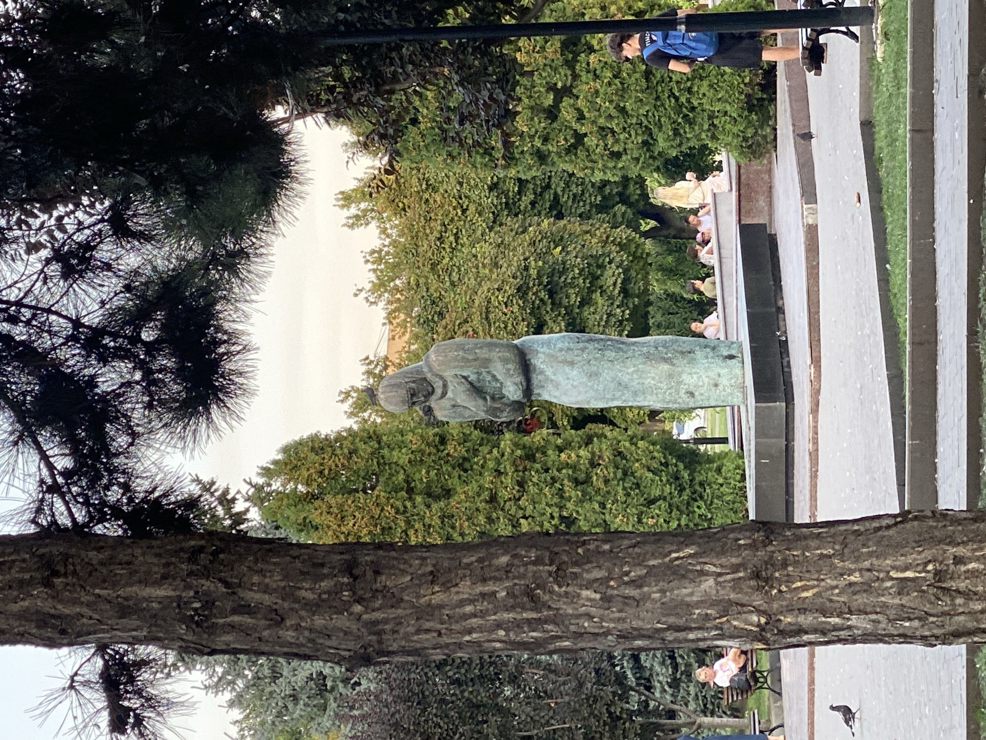 The image shows a statue of a woman holding a child, located in a park. The statue is made of bronze and is weathered green. It is set on a stone pedestal. In the background, there are several trees and bushes, as well as a few people sitting on benches. A young man with a backpack is standing to the right of the frame. A pigeon is visible on the ground in the foreground. The scene appears to be in a public park in Chișinău, Moldova, likely near the "Laiz" monument dedicated to the victims of the 1941 deportations.