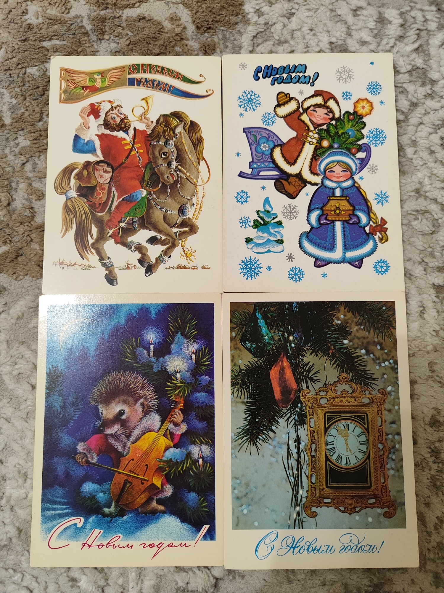 The image displays four vintage New Year's greeting cards arranged in a 2x2 grid. The cards are resting on a textured, carpeted surface.
The top-left card features an illustration of a man with a beard, wearing a fur-trimmed red coat and a hat, riding a horse. He is holding a horn to his lips, and a banner with a bird motif and Cyrillic text is unfurled above him. The bottom of this card depicts a snowy village scene.
The top-right card shows two children celebrating the New Year. One child, dressed in a red fur coat, sits in a sled pulled by a reindeer, holding a Christmas tree adorned with ornaments. Another child, in a blue fur coat and hat, stands below holding a gift box. The background is decorated with snowflakes. The text on this card reads "С Новым годом!" (Happy New Year!).
The bottom-left card depicts a hedgehog dressed in a red coat playing a cello. The hedgehog is positioned in a snowy, dark blue forest setting with lit candles on a Christmas tree. The text at the bottom reads "С Новым годом!" (Happy New Year!).
The bottom-right card features a close-up of a Christmas tree branch with an ornate, gold-framed clock hanging from it. The clock face shows roman numerals. The background is a blurry, snowy scene, and the text at the bottom reads "С Новым годом!" (Happy New Year!).
There is no indication of the city of Pavlodar, Kazakhstan, in this image.