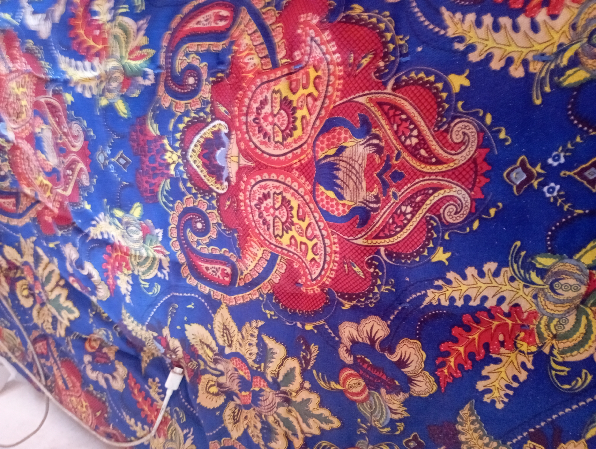 The image displays a close-up view of a blue textile with a paisley pattern. The pattern features swirls and stylized floral motifs in red, yellow, and green. A white USB cable is visible in the upper left corner, partially resting on the fabric. The lighting suggests an indoor environment. No specific location in Vostochnoe, Kyrgyzstan, can be identified from the provided image.