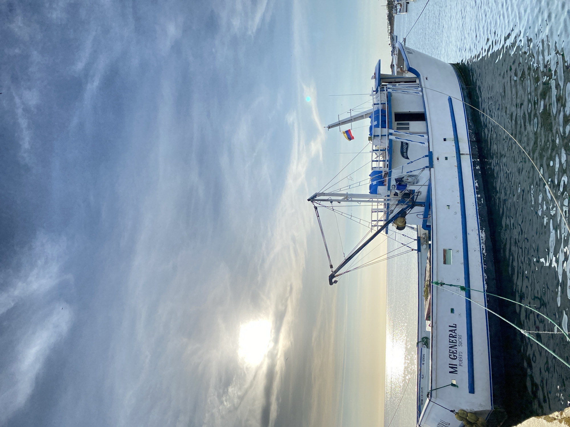 The image displays a large fishing boat, predominantly white with blue trim, docked in calm water. The boat, named "MI GENERAL," is seen from its starboard side, secured by mooring lines to an unseen dock. Additional text on the hull includes "PUERTO SUCRE," "AN-CA-0091," and "ALIX."
A mast with rigging and a boom is visible towards the front of the boat, indicating its function as a fishing vessel. A flag featuring horizontal yellow, blue, and red stripes, consistent with the Venezuelan flag, flies from the mast. The setting is likely a harbor or marina in Juan Griego, Venezuela, given the flag and the provided context.
The scene is illuminated by a low-lying sun, which creates a bright glare on the water's surface, suggesting either early morning or late afternoon. The sky is partly cloudy, with wispy, scattered clouds across a pale blue expanse. The horizon is visible in the distance, where the tranquil water meets the sky. There are no people visible in the frame, contributing to a peaceful and serene atmosphere at the port.