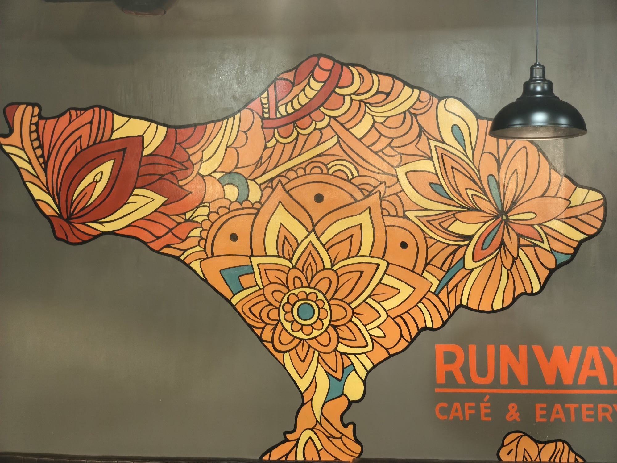 The media file is a photograph of a mural on a dark gray wall, likely inside a building given the presence of a black pendant light fixture hanging from the ceiling. The mural is a stylized representation of the island of Bali, rendered in a vibrant, mandala-like pattern. The colors used are predominantly shades of orange, yellow, and red, with accents of teal. The design features intricate floral motifs and abstract shapes, creating a visually engaging and decorative piece.
In the lower right portion of the mural, there is visible text that reads "RUNWAY" in large orange lettering. Below this, in smaller orange lettering, it says "CAFÉ & EATERY". This indicates that the mural is likely part of the branding or decoration for a café or eatery named Runway.
The scene is static, focusing on the artwork and the immediate surroundings. There are no people visible in the frame. The lighting appears to be artificial, likely from the pendant light and possibly other ambient lighting within the establishment. The overall impression is one of a modern, artistic interior design. The location cues, specifically the text "RUNWAY CAFÉ & EATERY," strongly suggest the setting is a commercial establishment. Considering the context of Kuta, Indonesia, the mural's style could be inspired by local artistic traditions or a general aesthetic that appeals to tourists and locals alike.