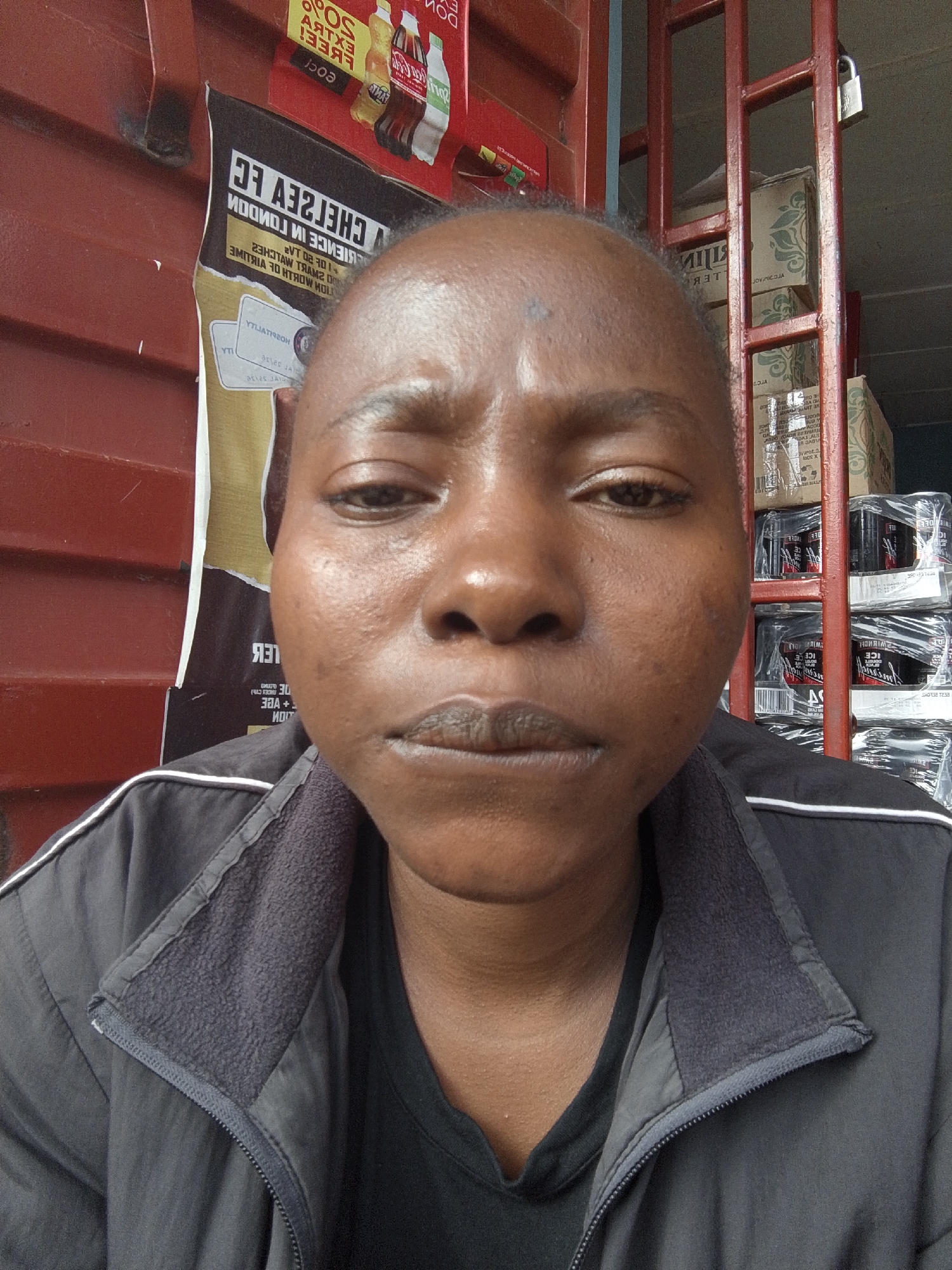 A close-up shot of a dark-skinned person, likely female, wearing a black jacket. The person is indoors, standing in front of a maroon corrugated wall and a metal grid structure. Behind the structure, there are stacked boxes of various beverages, including those labeled "ICE." A partially visible advertisement with "CHELSEA FC" and "LONDON" printed on it is also present on the wall. The location of the image is likely a small shop or kiosk, potentially in Jos, Nigeria. The focus is on the person's face.
