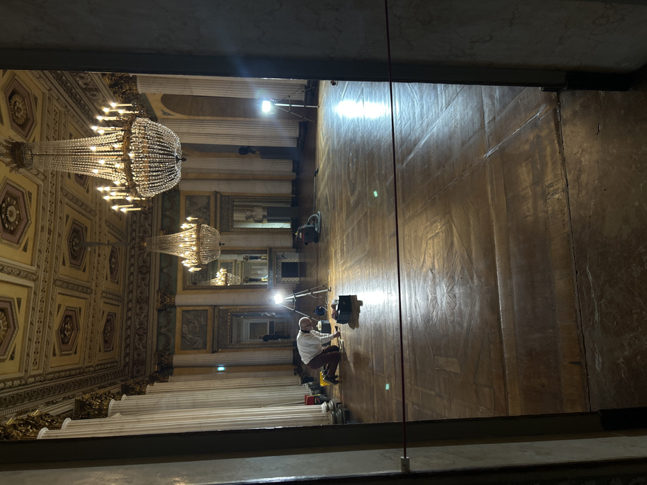 The image depicts a grand hall within the Palazzo Clerici in Milan, Italy, viewed through a doorway. The scene captures ongoing maintenance or preparation activity.
**Environment:** The hall features a high, elaborate coffered ceiling adorned with gilded moldings and painted panels in geometric patterns of gold, cream, and muted tones. The walls are lined with tall, fluted Corinthian columns in light marble, supporting a decorative entablature. Between the columns, large arched mirrors reflect the room's interior, including a third crystal chandelier. Below the entablature and above the mirrors, painted friezes or relief panels with classical motifs are visible. A decorative fireplace mantelpiece with a white classical bust sculpture sits in the far background, with another dark bust on a pedestal further to the right. The floor is made of polished, aged parquet wood with intricate geometric inlay patterns, including star-like central medallions.
**Lighting:** Two large, multi-tiered crystal chandeliers with multiple candle-style bulbs hang from the ceiling, one prominent in the mid-right, the other further back. Additionally, two bright, professional-grade LED or floodlights on tripods are positioned on the floor, illuminating specific areas and casting strong reflections on the polished parquet.
**People:** One male individual, with a beard and wearing a light-colored long-sleeved shirt and dark trousers, is kneeling on the floor in the left-midground. He is focused on an area directly in front of him, actively engaged in a task with his hands near small containers and a dark, rectangular object (possibly a toolbox). A second, less distinct figure is partially visible standing further back in the hall near a far doorway on the left.
**Objects:** Near the kneeling individual, several items are on the floor: a red container, other dark containers, and a dark, rectangular case. To his right, an industrial vacuum cleaner with a hose is present. Two portable floodlights on tripods are positioned in the midground, contributing significant artificial illumination. A red cord or rope is strung horizontally across the lower middle of the frame, attached to the doorframes, acting as a barrier.