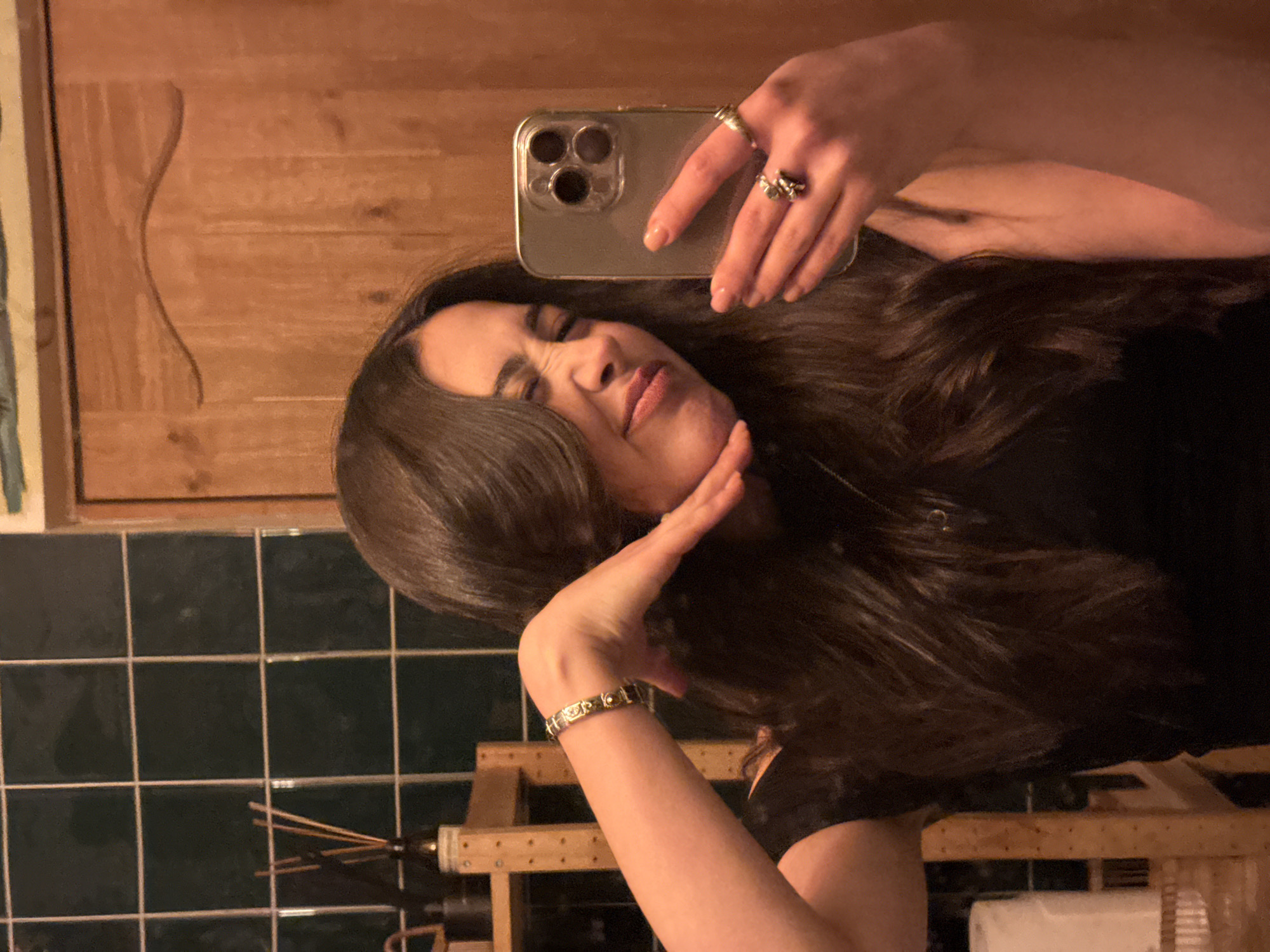 A woman is taking a selfie in a bathroom with green tiles. She is holding an iPhone in her right hand and her left hand is near her face. She is wearing rings on both hands and a bracelet on her left wrist. A wooden cabinet and a diffuser are visible in the background. There is no evidence to indicate the location is in Yerevan, Armenia.