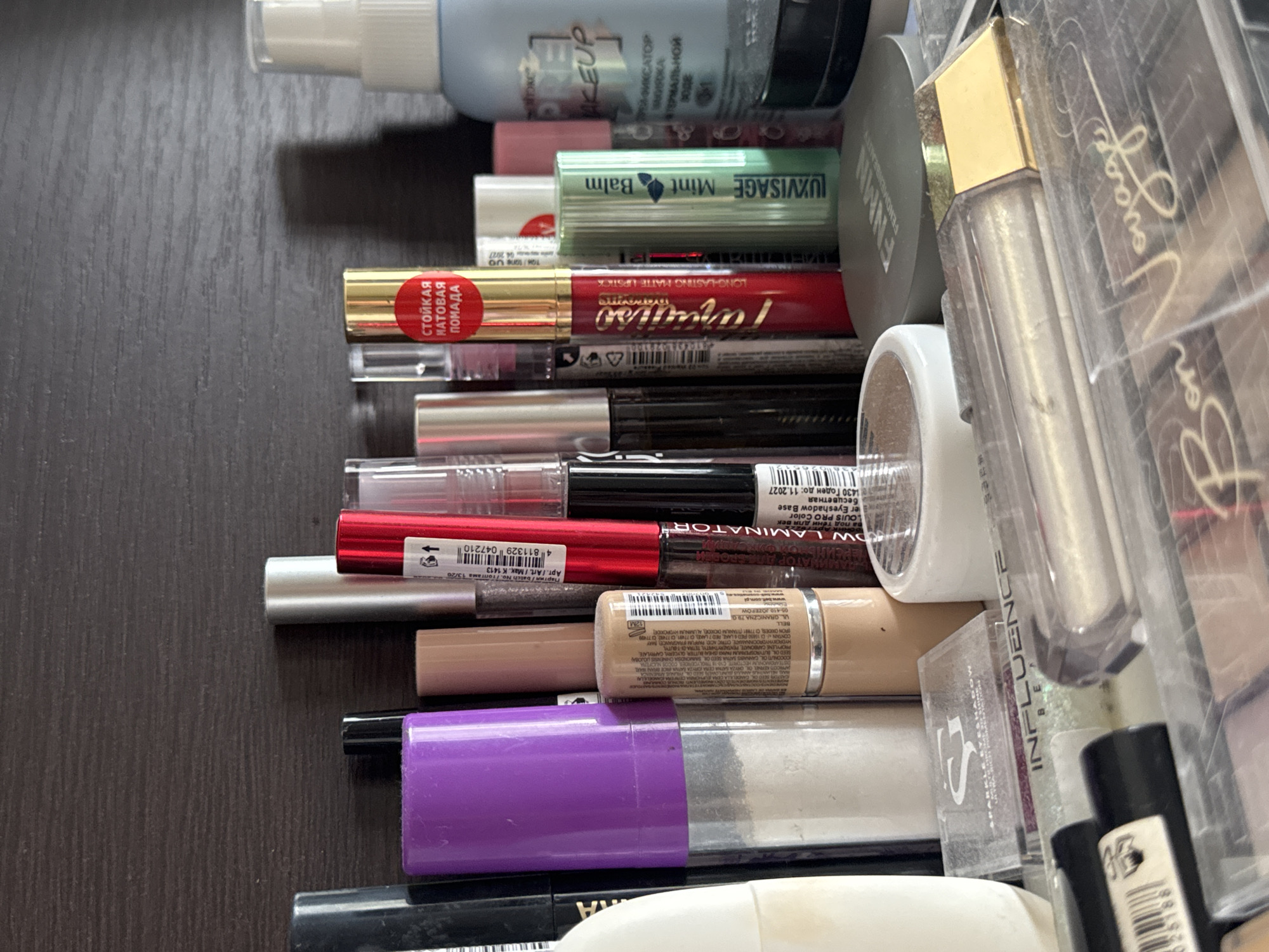 This is a close-up, eye-level, medium shot of a shelf filled with various makeup products. The background is a dark, textured surface, likely a wooden shelf. The lighting is overhead and creates shadows. The makeup items are densely packed, with some out of focus due to the shallow depth of field.
In the foreground, a clear plastic container with a gold lid is prominent, containing a shimmery liquid product, possibly lip gloss. To the left and slightly behind it, another clear plastic container holds multiple shades of eyeshadow or blush, with the text "Bon Voyage" visible.
Further back, various lipsticks, lip glosses, and makeup tools are visible. Notable items include a bright red lipstick with gold packaging and Russian text that translates to "Long-lasting matte lipstick," and a green tube labeled "LUXVISAGE Mint Balm." There are also several tubes of mascara or liquid eyeliner, one in black and another with a silver cap. A cylindrical purple container, possibly for powder, stands out.
The overall impression is a personal collection of makeup, neatly but densely arranged on a shelf. There are no people or actions depicted, only a static arrangement of objects. The specific location of Яромінскі сельскі Савет, Belarus, cannot be determined from the visual information alone.