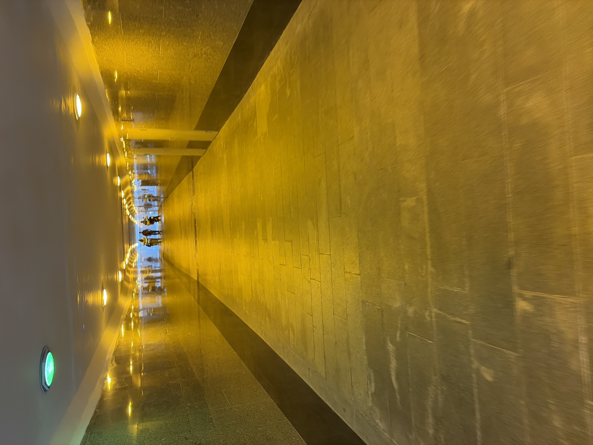 A vertical, eye-level shot captures a brightly lit pedestrian underpass. The walls of the underpass are clad in dark, speckled tile, with a horizontal band of dark material separating the tiled lower section from the white ceiling. Rows of yellow lights are embedded in the ceiling, casting a warm glow and creating long reflections on the wet, stone-paved floor. A single green light is visible on the ceiling near the viewer. In the distance, a group of three individuals, silhouetted and indistinct, are walking away from the camera. The scene is clean and modern, suggesting an urban environment. The overall impression is one of a safe and well-maintained public space, likely in Moscow, Russia.