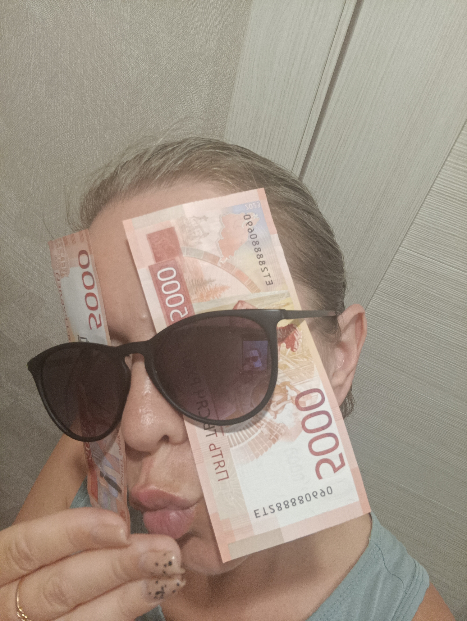 A woman poses with sunglasses and 2000 Russian ruble banknotes. The banknotes are draped over the face, one held in the hand and two tucked under the sunglasses. The background includes a textured wall and a paneled wall, possibly indoors. The photo was taken in the city of Chaikovsky, Russia.