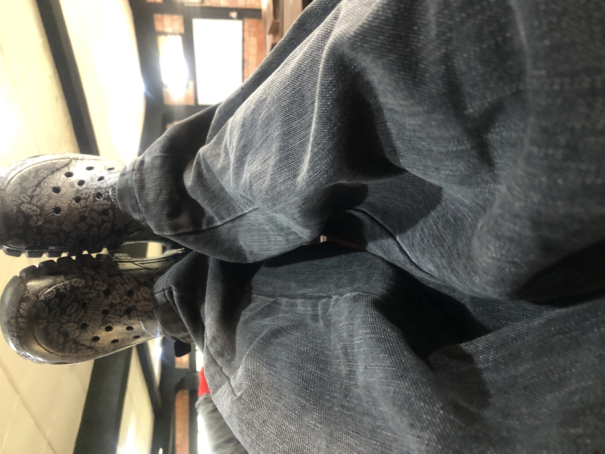 The image is a close-up, low-angle shot of a person's legs wearing dark denim jeans and patterned black Crocs. The perspective is from the person's own point of view, looking down at their legs and feet. The Crocs have a detailed floral or leaf-like pattern in a lighter shade. The jeans are wrinkled and folded, obscuring much of the lower body. The background is out of focus, showing what appears to be an indoor environment with lights and brickwork, possibly a cafe or waiting area. No other people are visible in the frame. The overall lighting is warm, likely from overhead lights.