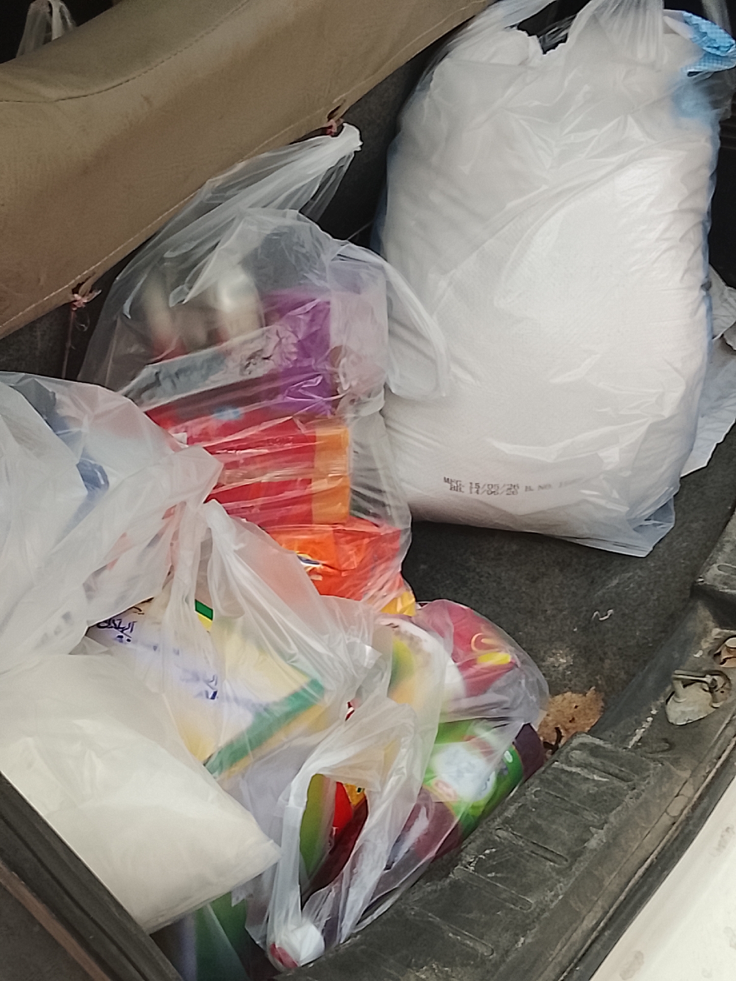 The image depicts the contents of a car trunk. Visible inside the trunk are several clear plastic bags filled with various items, including what appears to be food products and possibly household goods. A large bag of white powder, likely sugar or flour, is prominent in the back. The trunk interior is dark and shows signs of wear and tear. The city and colony mentioned are not discernible from the image. There are no people visible.
