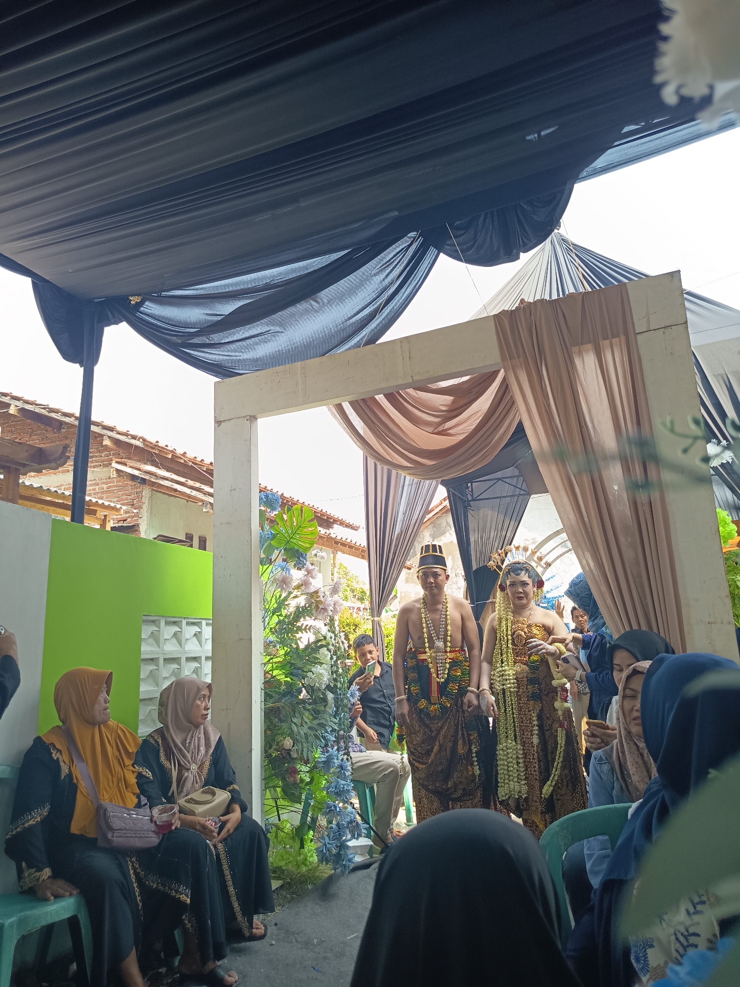 In an outdoor setting, likely a wedding reception, a young couple is standing under a decorated archway. The man is wearing traditional attire, including a headdress and necklaces, while the woman is adorned in a gold-colored dress and a crown. They are surrounded by guests, some of whom are seated and others standing. The environment is covered by a black tent or canopy, with a light-colored fabric draping from the archway. In the background, there are buildings with tiled roofs and some greenery. The overall atmosphere appears to be festive and celebratory.