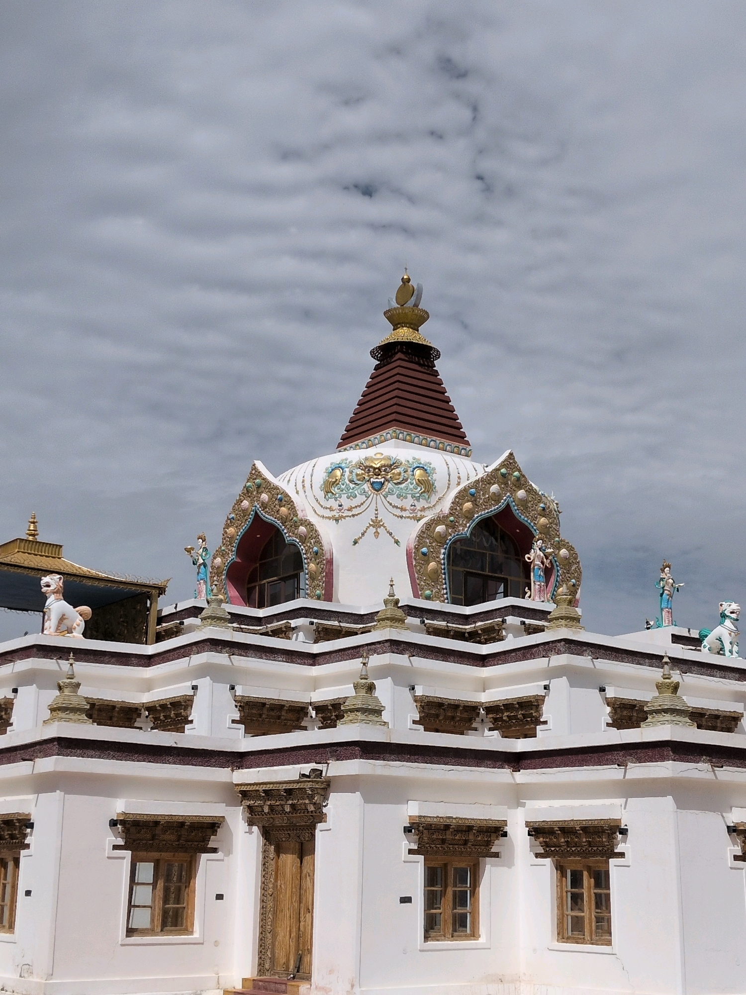 A white Buddhist stupa with ornate gold and blue detailing stands against a cloudy sky in Martselang, India. The stupa is multi-tiered, with a conical roof topped by a golden spire. The base of the stupa features several decorative windows with carved wooden frames, and small stupas adorn the edges of the upper levels. Two white lion-like statues are positioned on the roof, along with two small human figurines dressed in blue and red. The lower part of the image shows a white building with similar wooden-framed windows and a large wooden door. The sky is filled with a layer of grey clouds.
