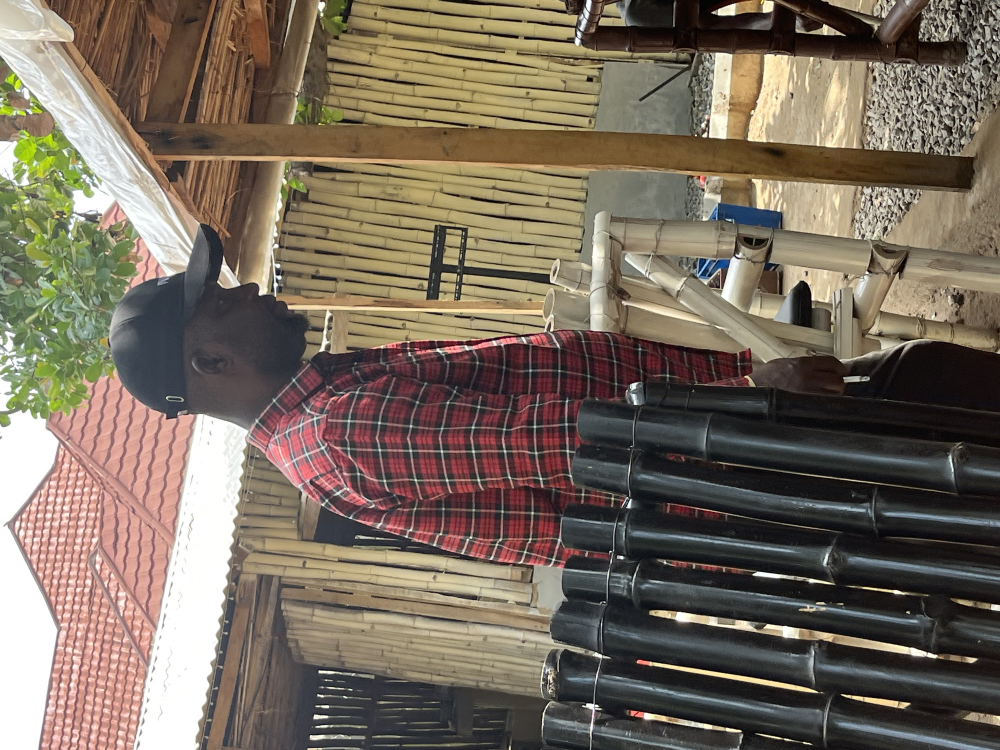 The image depicts a man wearing a black baseball cap and a red and black plaid shirt, standing outdoors. He is positioned in front of a structure made of bamboo poles, with a corrugated metal roof and a red tiled roof visible behind it. To the man's left, there is a dark-colored fence or barrier made of vertical poles. In front of him and to his right are various pieces of furniture and structures also constructed from bamboo. The ground appears to be covered in gravel and dirt. A blue trash bin is visible in the background. The scene suggests an outdoor seating area or a construction site, possibly a market stall or a restaurant with a natural, rustic aesthetic. The lighting indicates it is daytime.