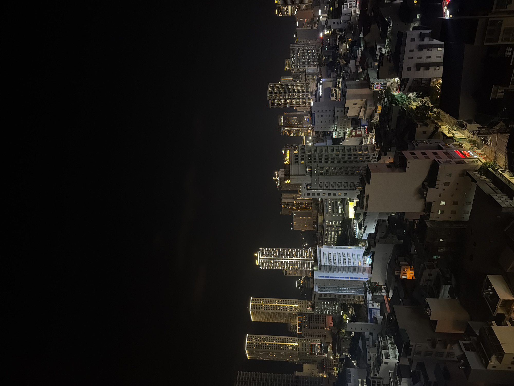 A nighttime view of Nha Trang Ward, Vietnam, shows a city skyline under a dark sky. Illuminated buildings of varying heights and architectural styles are on the right. The left side of the image is dominated by black space. Lights from buildings, streetlights, and traffic can be seen.