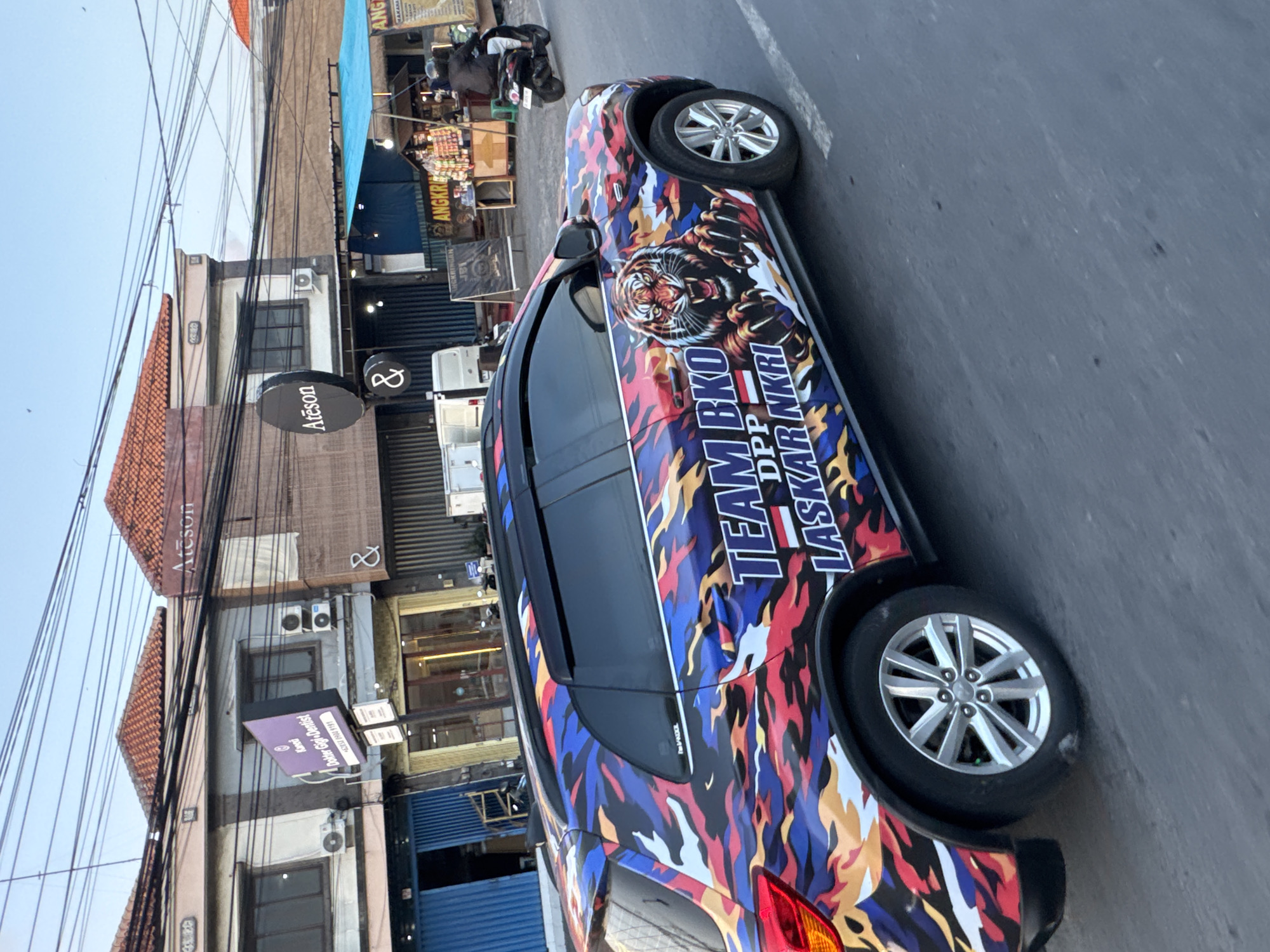 The image shows a colorful car wrapped in a camouflage design with a tiger graphic and text on its side, driving on a street in Denpasar, Indonesia. The text on the car reads "TEAM BKO DPP LASKAR NKRI." In the background, there are buildings with shops and signs, including one that says "Ateson" and another that appears to be for a dentist. A person is riding a motorcycle in the background. The sky is clear and blue.