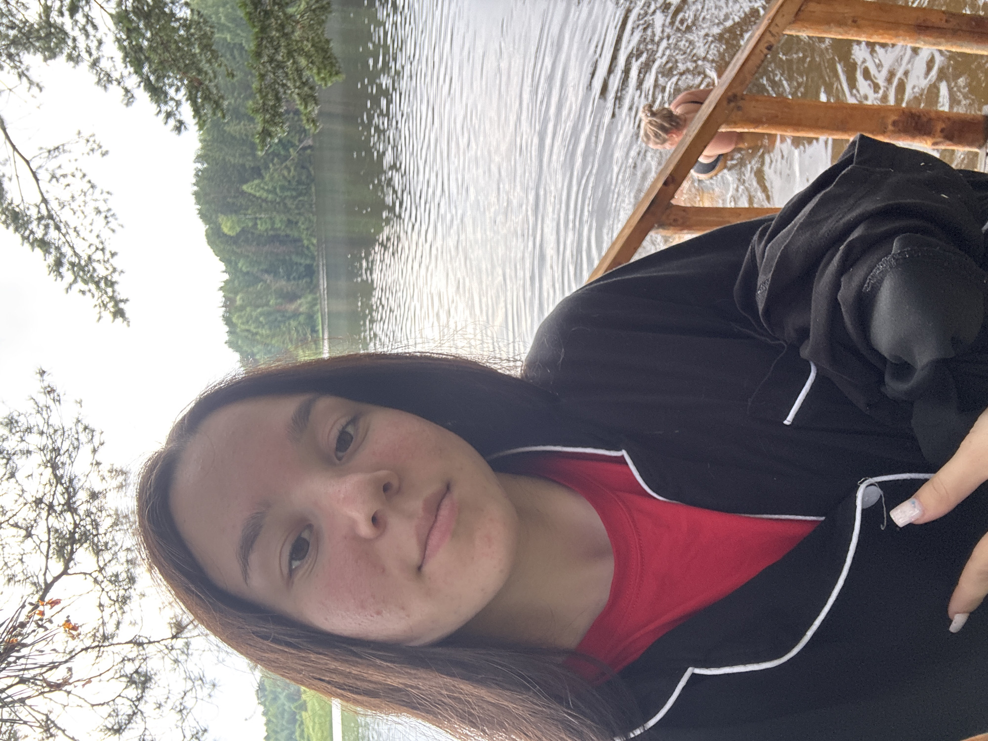 A young woman with long brown hair and light skin takes a selfie by a lake in Pozdyshki, Russia. She is wearing a red t-shirt and a black robe with white trim. In the background, two other people are swimming in the lake, and a wooden dock is visible. The lake is surrounded by trees, and the sky is overcast. The water is calm and reflects the trees. The overall atmosphere is relaxed and natural.