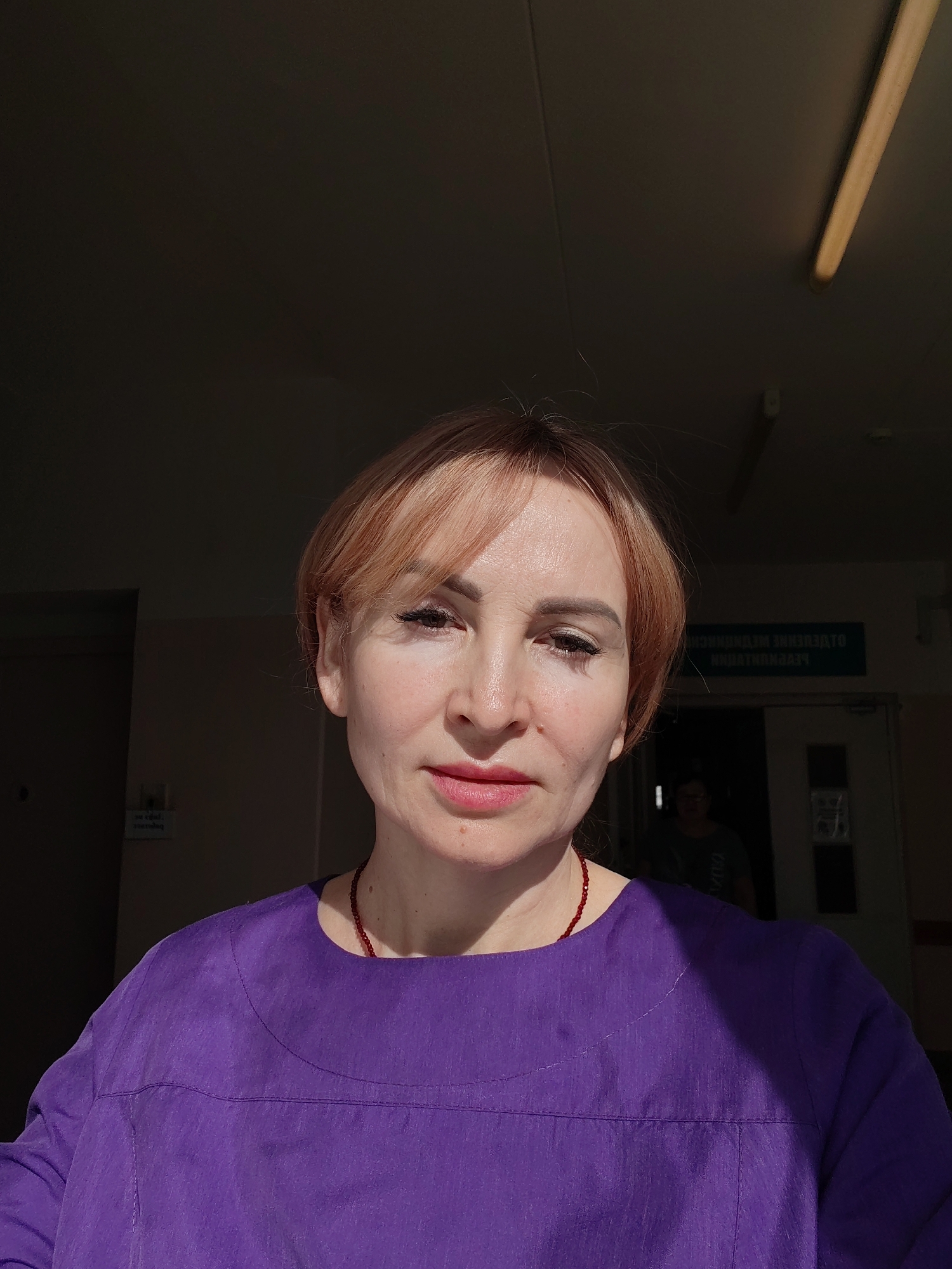 A middle-aged woman with short, light brown hair stands in a dimly lit interior, wearing a purple top and a red beaded necklace. Sunlight illuminates part of her face. Behind her is a door with a sign in Cyrillic, a second person is slightly visible through the open doorway. The location is potentially in Chaikovsky, Russia, judging by the Cyrillic text. A fluorescent light fixture is visible on the ceiling. The scene appears to be indoors.