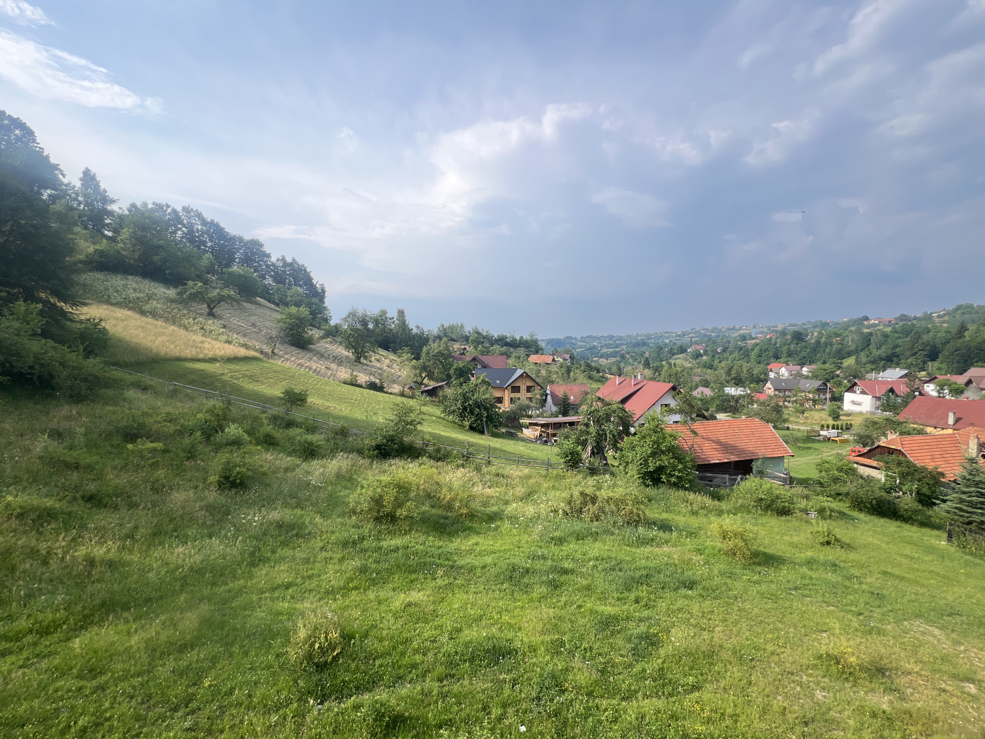This media file captures a serene, bucolic scene in a rural village nestled amidst rolling green hills. The foreground is dominated by a lush, overgrown meadow with tall grasses and scattered wildflowers, indicative of a warm season. A wooden fence runs along the edge of the meadow, separating it from a terraced hillside.
Further into the scene, a cluster of houses with red-tiled roofs are visible, scattered amongst the trees. Some houses appear to be traditional wooden structures, while others are more modern. The architecture suggests a European or Eastern European setting. The houses are surrounded by dense foliage, with mature trees providing shade and framing the village.
The sky above is dynamic, with a mix of bright sunlight breaking through scattered clouds. This suggests it might be late morning or early afternoon, with potential for changing weather. The overall atmosphere is peaceful and tranquil, evoking a sense of rural simplicity and natural beauty. There is no discernible activity or event taking place; the image appears to be a static landscape photograph. No text is visible in the media file.