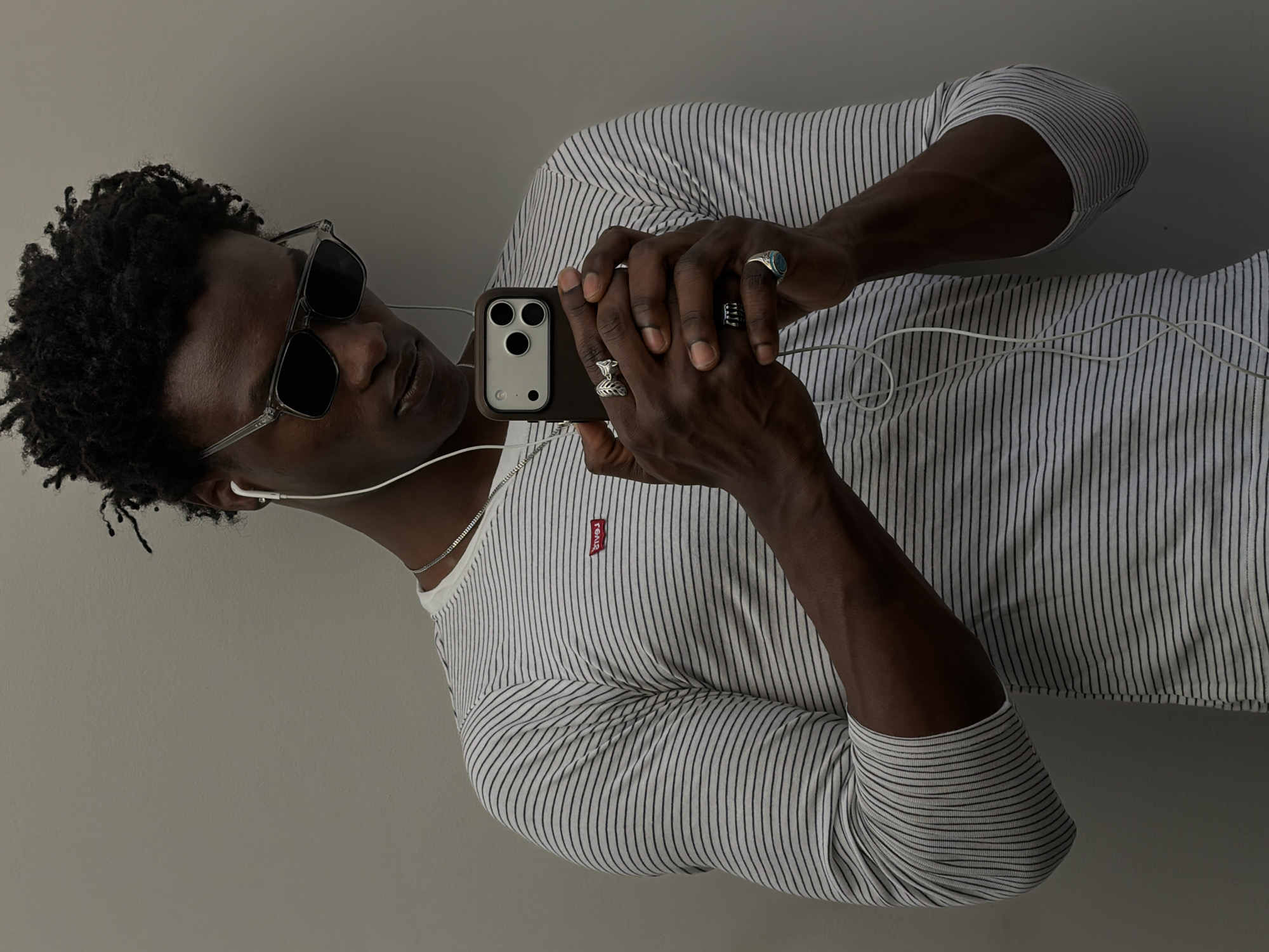 A man is shown from the chest up, holding his cell phone in front of him as if taking a selfie. He has dark skin and short curly hair. He is wearing sunglasses, earbuds, a white t-shirt with narrow black stripes, a silver chain necklace, and multiple rings. He is holding a dark cell phone. The background is a plain, light gray wall. No information suggests this image was taken in London, United Kingdom.