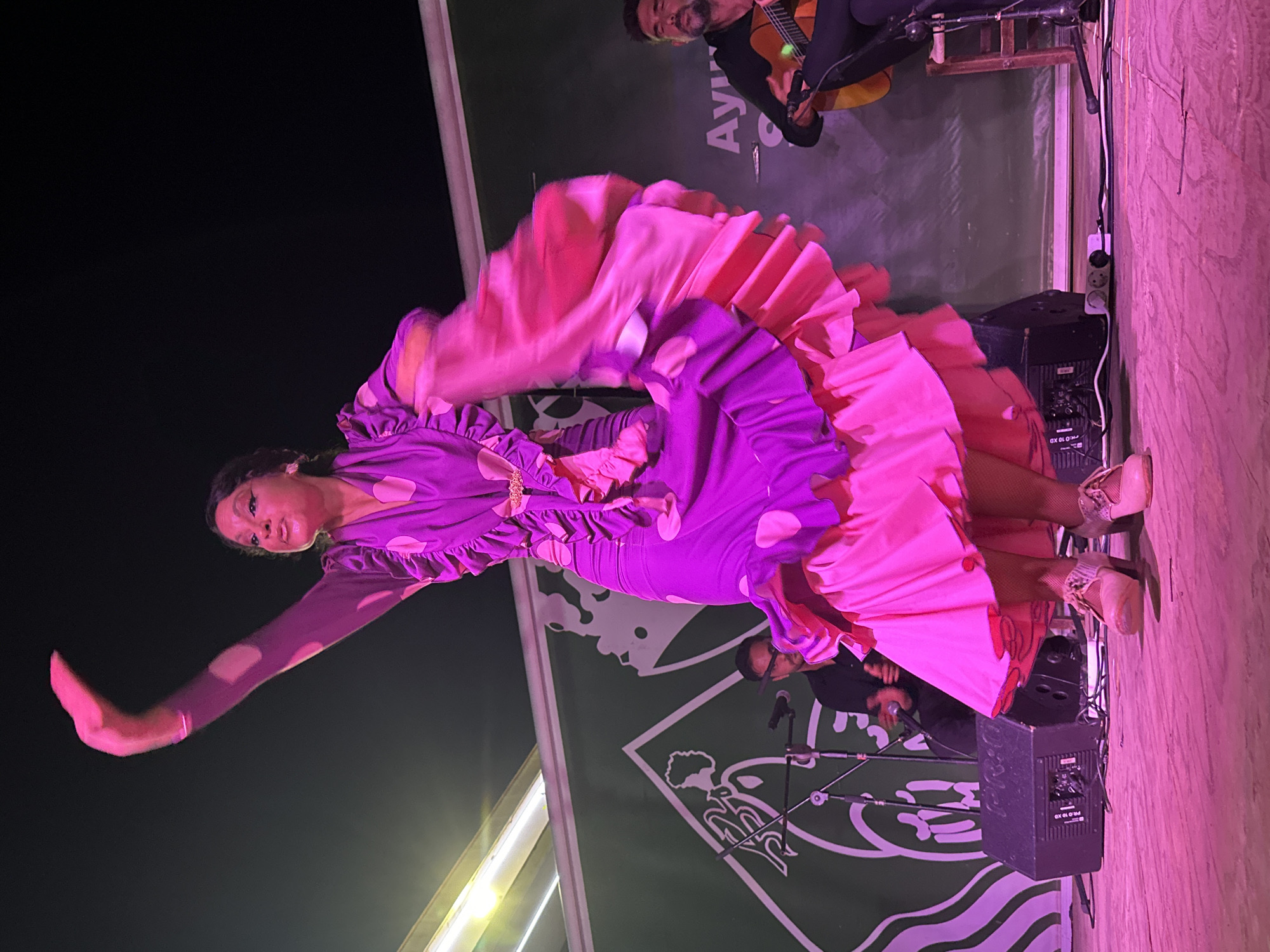 In San José Del Valle, Spain, a flamenco dancer in a vibrant purple dress with large pink polka dots performs on a stage. The dress features ruffled sleeves and a tiered, flowing skirt in shades of pink and purple. Her right arm is raised in a dramatic gesture, and her left hand is partially obscured by the skirt. She wears light-colored, heeled shoes and fishnet stockings.
Behind the dancer, two musicians are visible. On the left, a man in a black shirt plays a guitar, his head tilted down as he concentrates. A microphone stands in front of him. On the right, another man, also in dark clothing, plays a guitar and sings, his face visible and eyes closed.
The stage is made of wood and is illuminated by pink lighting. In the background, a large green banner with white graphic elements is stretched across the scene. Two black speaker boxes are positioned on the stage floor. Electrical cords and a power strip are visible in the foreground, connecting to the speakers. The overall atmosphere suggests an outdoor evening performance.