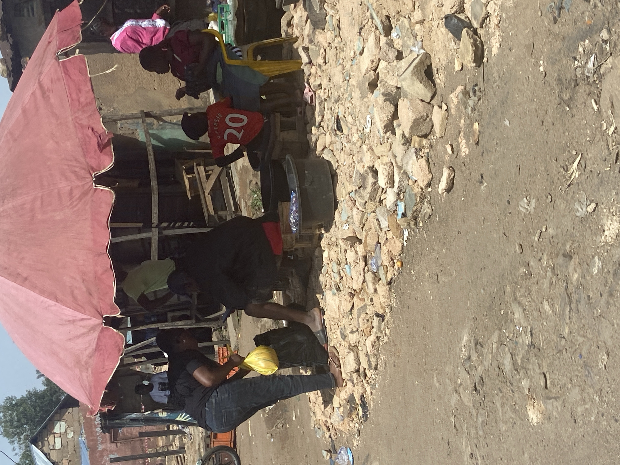 The image shows a street scene in Bukuru, Nigeria, likely during the day. A large, pink umbrella or canopy shades a market stall. Underneath the canopy, several people are visible. In the foreground, a person wearing a black t-shirt and blue jeans is holding a yellow bag. To their right, another person in a black outfit is also present, and further back, a person in a red shirt with the number "20" and the name "VAN PERSIE" is visible. The background is a rough, uneven surface covered in rocks, debris, and some vegetation. The overall setting appears to be a busy, informal marketplace with simple structures and a dusty environment. The lighting suggests it is daytime.