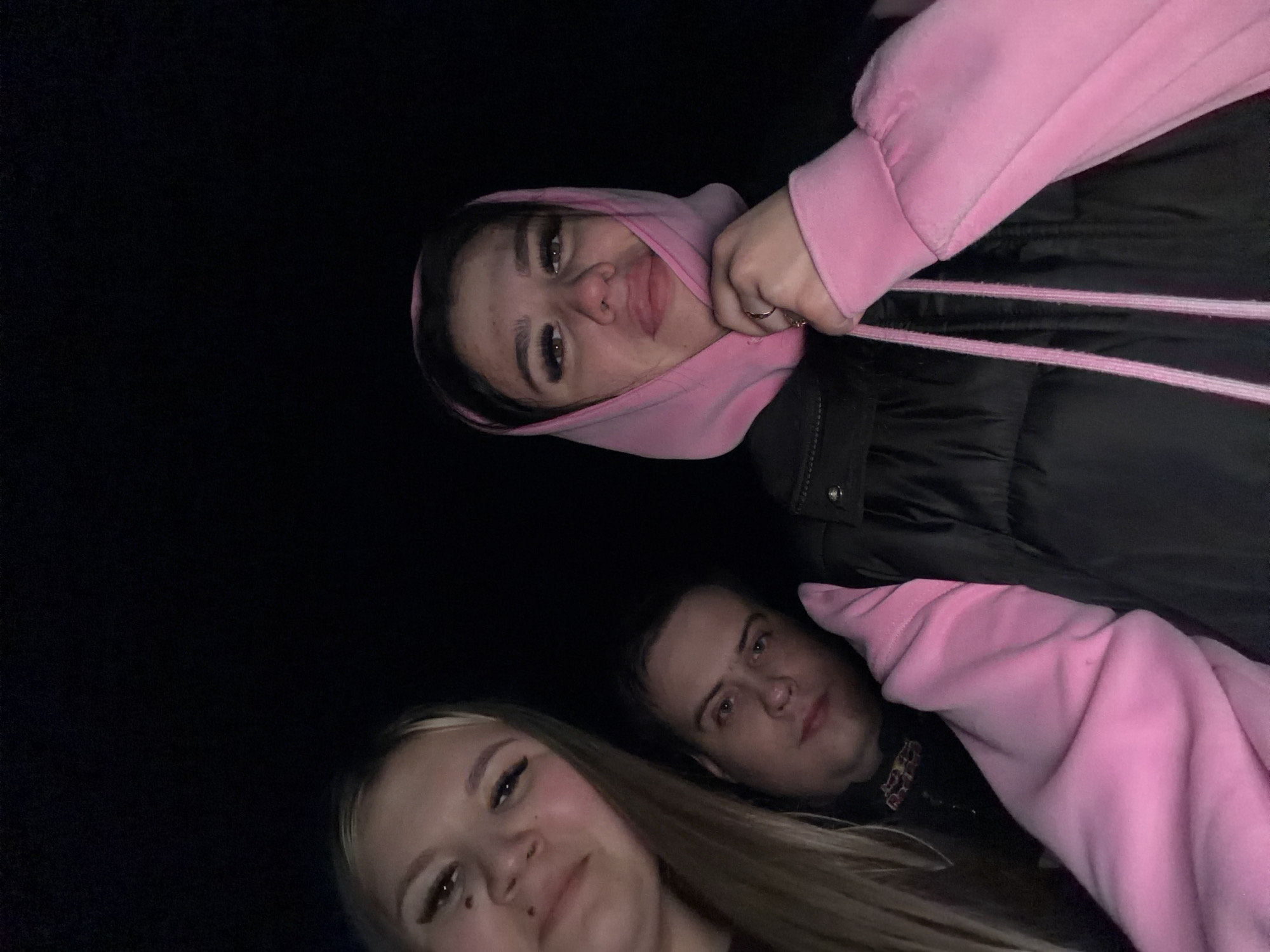Three individuals are captured in a low-light photograph. The background is uniformly dark. A woman wearing a pink hooded sweatshirt and a black puffer jacket is positioned at the top of the frame. A woman with blonde hair is at the bottom left, and a man is situated towards the bottom center. The location in the city of Фоки, Russia, is not discernible from the image.