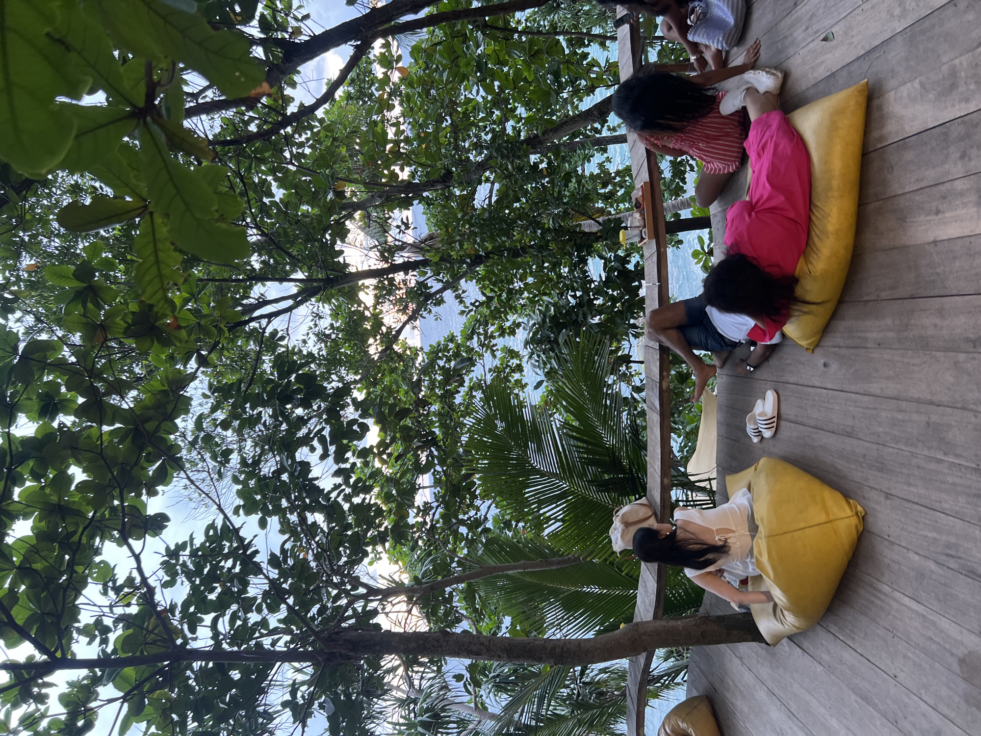 Four people are relaxing on a wooden deck overlooking a tropical ocean view. Two women are seated on large, yellow beanbag chairs. One woman is wearing a pink and white striped top and a pink skirt. She is leaning back against a beanbag chair. The other woman is wearing a white top and a light-colored skirt. She is sitting with her legs extended on another beanbag chair. Two other people, one woman and one man, are partially visible in the background, also seated on the deck.
The scene is set amidst lush green trees and palm fronds, suggesting a tropical location. The ocean is visible in the background, with calm, blue water. The lighting suggests it is either early morning or late afternoon, with soft sunlight filtering through the leaves. The mood appears relaxed and leisurely. There are a pair of white and black striped sandals on the wooden deck. No visible text is present in the image. The location context provided is Karon, Thailand.