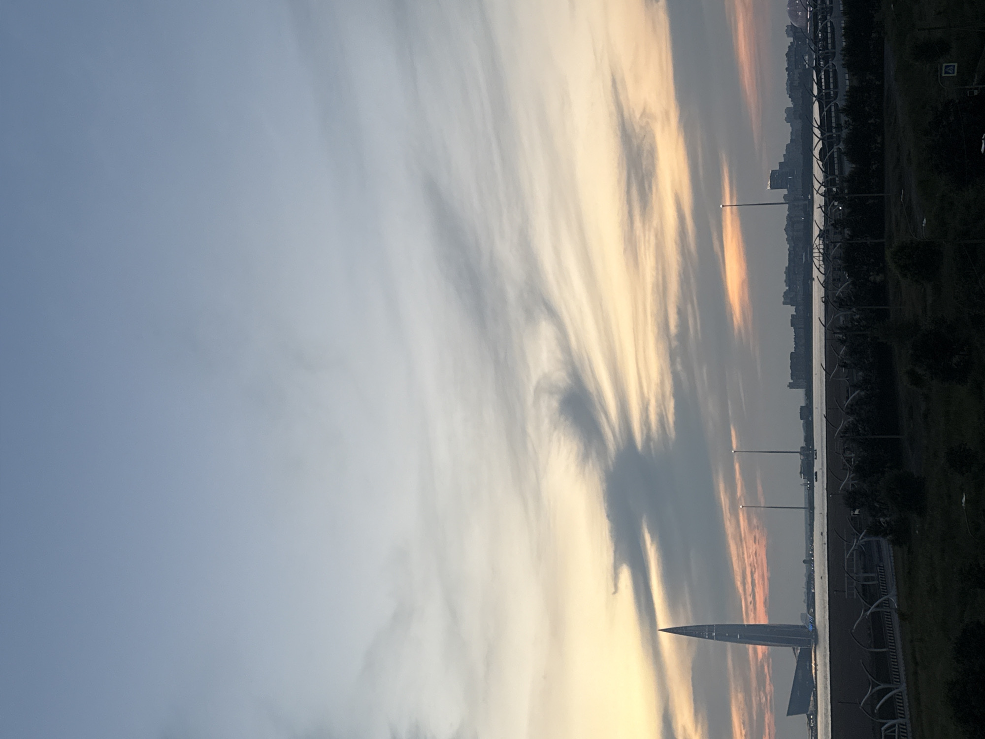 The image depicts a cityscape at dusk, with a prominent modern skyscraper on the left side. The skyscraper, which is likely the Lakhta Center in Saint Petersburg, Russia, is illuminated with blue lights at its base. It stands against a sky streaked with orange and grey clouds, indicating the setting sun. In the background, a body of water separates the foreground from a distant city skyline composed of various buildings. A network of white, curved structures, resembling modern road barriers or architectural elements, runs across the middle ground. Trees and dark vegetation occupy the bottom portion of the image. Two tall, thin towers with lights at the top are visible in the distance, to the right of the main skyscraper. The overall atmosphere is serene and evokes the transition from day to night in an urban environment.