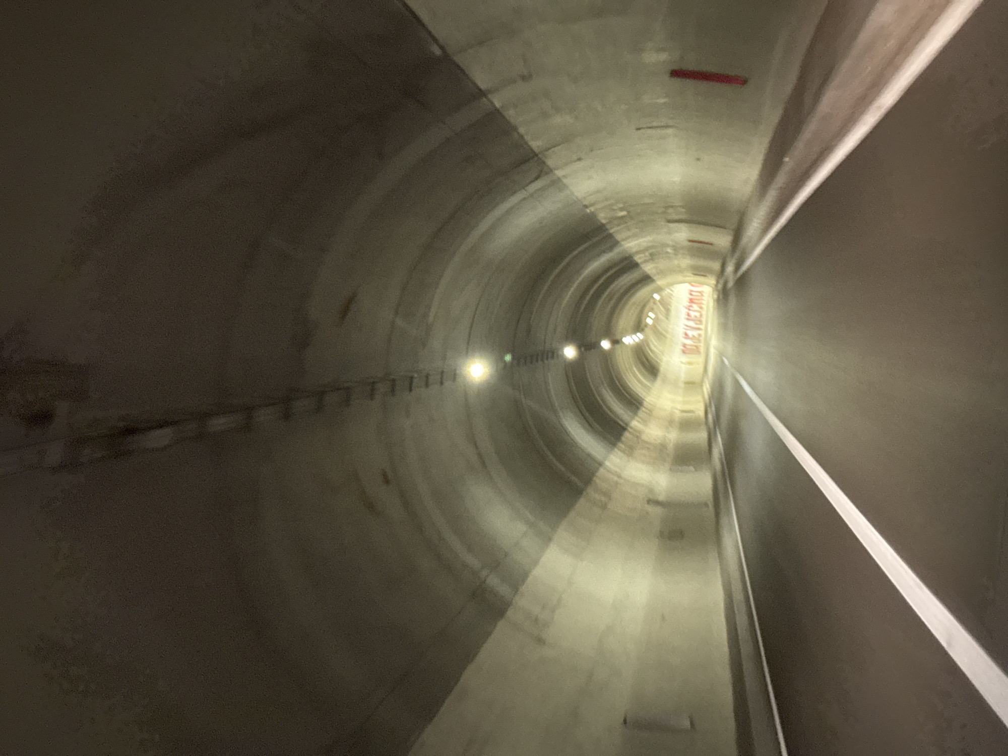 The image depicts the interior of a tunnel, likely a highway tunnel, as indicated by the asphalt road with white lane markings. The tunnel is illuminated by overhead lights, creating a sense of depth as they recede into the distance. The walls and ceiling of the tunnel appear to be constructed from concrete, with some textured or irregular surfaces visible. There is a red sign or banner visible on the far wall with text in what appears to be Cyrillic or Latin script. The perspective suggests the photo was taken from a moving vehicle. The location is identified as Bukovica, Montenegro, which is known for having several significant tunnels, including the Klisur and Vjetrenik tunnels which are part of the main road connecting Podgorica to the coast.