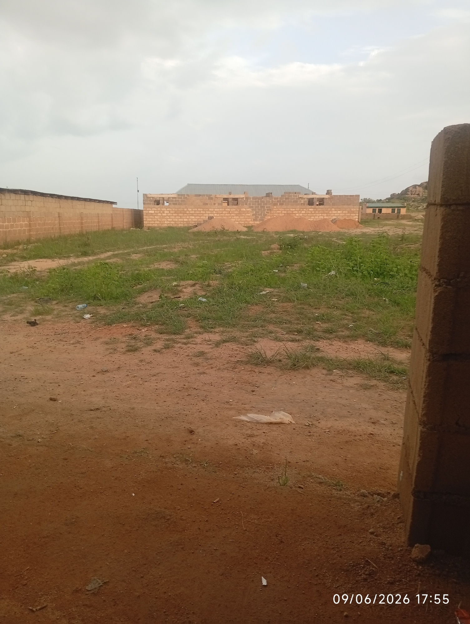 An exterior medium shot captures a construction site with a brick building under construction in the background. The building has a metal roof and appears to be in the early stages of construction, with unfinished walls made of concrete blocks. In the foreground, the ground is covered in dirt and patches of grass, with some scattered debris, including plastic bottles and wrappers. A large concrete pillar is visible on the right side of the frame. The sky is overcast with gray clouds. The timestamp "09/06/2026 17:55" is superimposed on the image. The location is described as Unknown, Unknown.