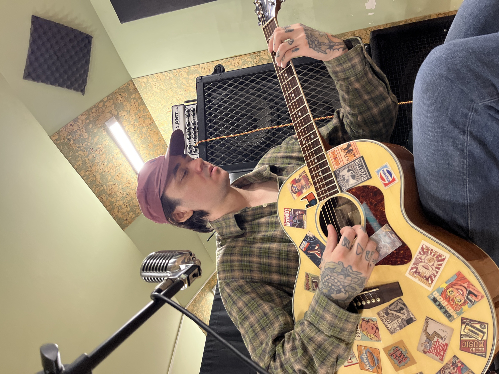 A person is playing a sticker-covered acoustic guitar in what appears to be a recording studio. The person, wearing a plaid shirt and a baseball cap, has tattoos on their hands. They are positioned near a microphone, suggesting they are recording their playing. The studio setting is evident from the presence of soundproofing panels on the wall and audio equipment in the background, including what looks like an amplifier.