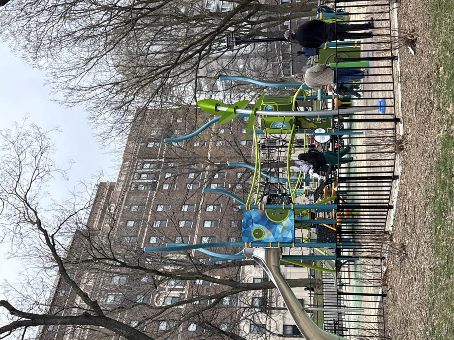 The image depicts a public playground in a park setting within New York City, United States of America, on an overcast day. The foreground shows mulched ground with sparse, bare shrubs, enclosed by a black metal picket fence. Dominating the midground is a modern playground structure featuring a large silver slide, green and teal vertical supports, black climbing nets, and a decorative panel with a blue and green geometric design. A tall, central playground element with green, leaf-shaped appendages is also present. Several individuals are within or immediately adjacent to the playground: one adult in a dark jacket and dark green pants assists a child on the climbing net, another adult in a light brown quilted jacket stands nearby with a small child, and a third adult in a black jacket stands with their back to the camera on the right. In the background, numerous deciduous trees, bare of leaves, frame a large, multi-story brick residential building with multiple windows, characteristic of New York City architecture. A street with vehicles and additional indistinct figures is visible beyond the playground.
