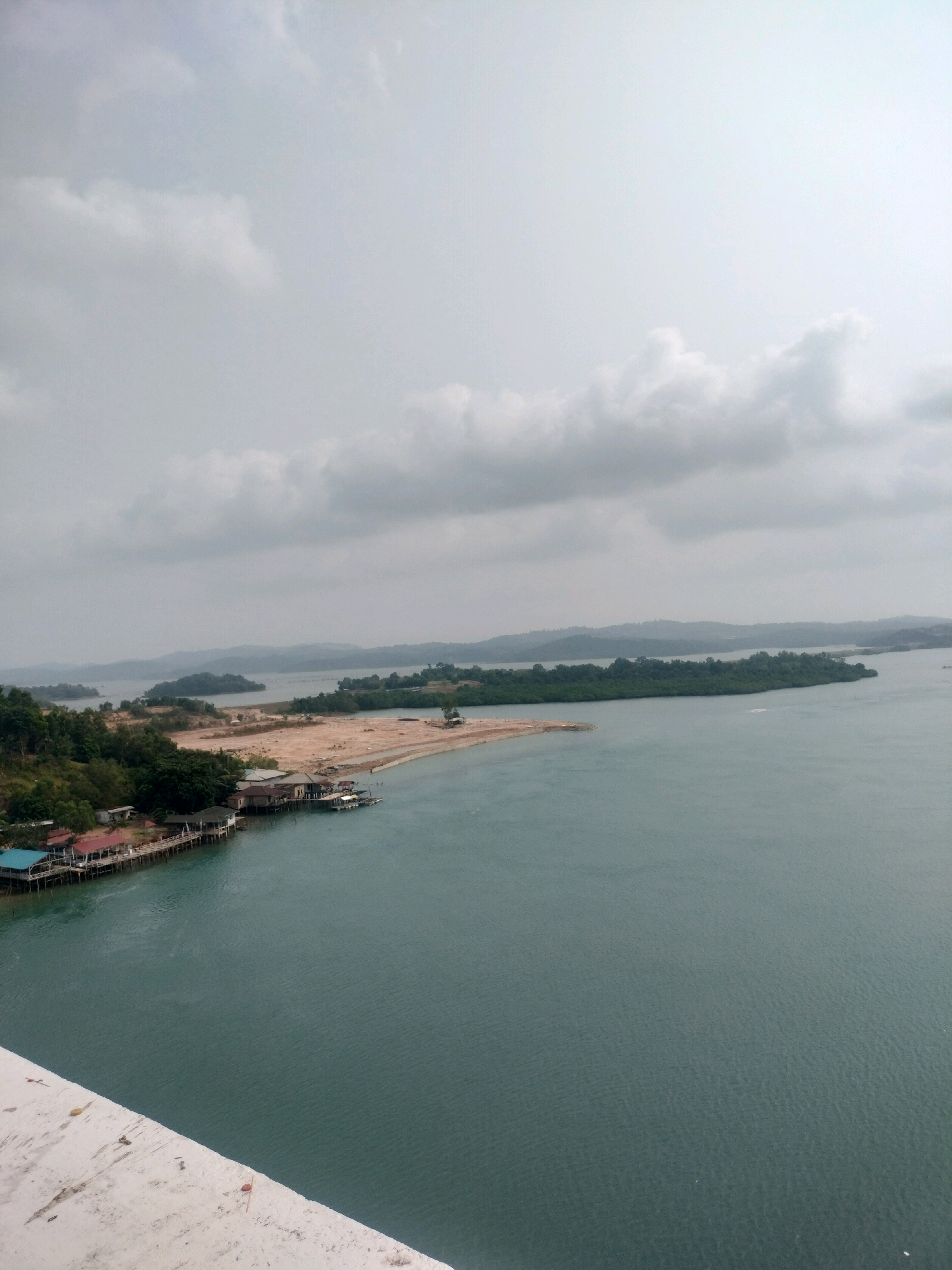 The image depicts a coastal landscape in Batam, Indonesia, likely from an elevated vantage point such as a bridge or building. The foreground shows a concrete structure with some scattered debris. The midground features a body of water, appearing as a bay or strait, with a distinctive teal color. Along the shoreline, there are several structures built on stilts, including houses with blue and red roofs, and a pier extending into the water. Some of these structures appear to be residential or possibly small businesses. Further inland, the land is covered with vegetation, with some areas appearing to be under development, showing exposed earth and construction materials. In the distance, a series of islands with green, tree-covered hills are visible, separated by the calm waters. The sky is overcast with diffused light, contributing to a muted color palette. The scene suggests a coastal settlement with ongoing development in a tropical environment.