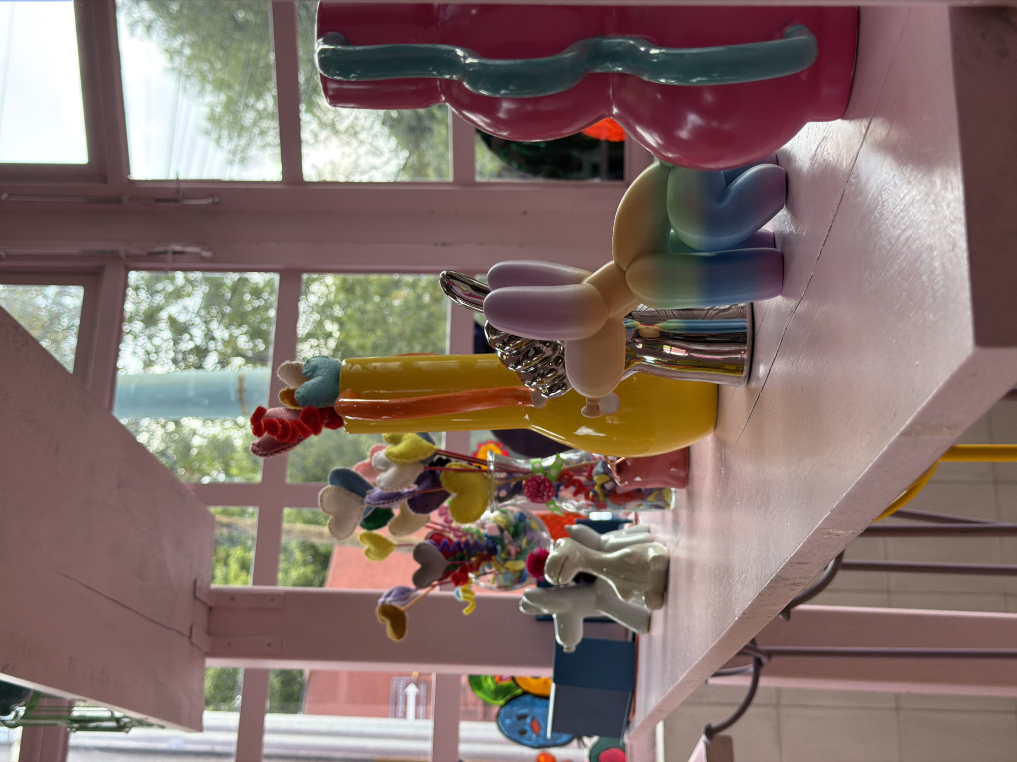 The image captures a whimsical interior scene in Puebla, Mexico. A shelf, painted a light pink, displays a collection of playful objects. These include a series of vases containing colorful felt hearts on sticks, a bright yellow vase, a balloon dog statue in pastel rainbow hues, and a bulbous pink statue with teal accents. The shelf is positioned in front of a window with multiple panes, offering a glimpse of the outside world, including buildings and greenery. The background is slightly out of focus. Below the shelf, glimpses of a staircase and other colorful decorations can be seen, suggesting a vibrant and playful environment. The image is slightly rotated, presenting an unusual perspective.