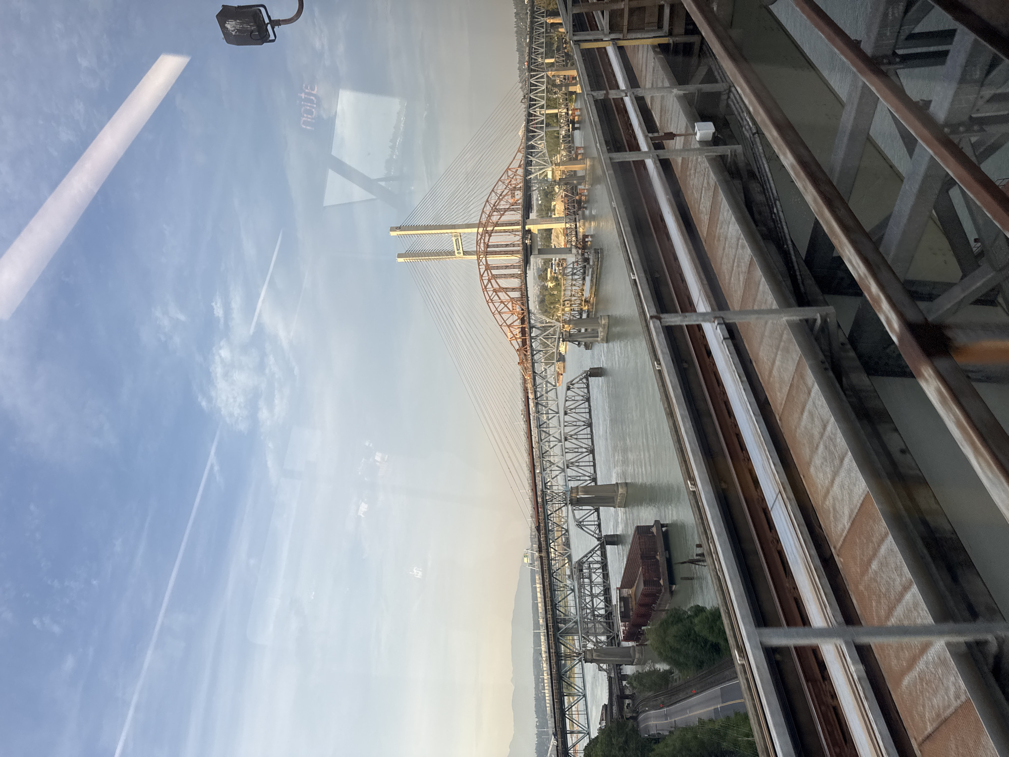This image captures a view of the Alex Fraser Bridge and the South Fraser Perimeter Road over the Fraser River in New Westminster, Canada, as seen from a passing train. The foreground features train tracks, with rust and metal structures indicating the perspective from within a train carriage. The middle ground displays the wide, gray expanse of the Fraser River, with barges docked along the bank. Several bridges are visible: an older truss bridge with a railway line, and a newer cable-stayed bridge, the Alex Fraser Bridge, distinguished by its white towers and orange arch. The background consists of a distant shoreline with trees and mountains under a hazy, pale blue sky. Sunlight reflects off the water and the train window, creating glares and obscuring some details.