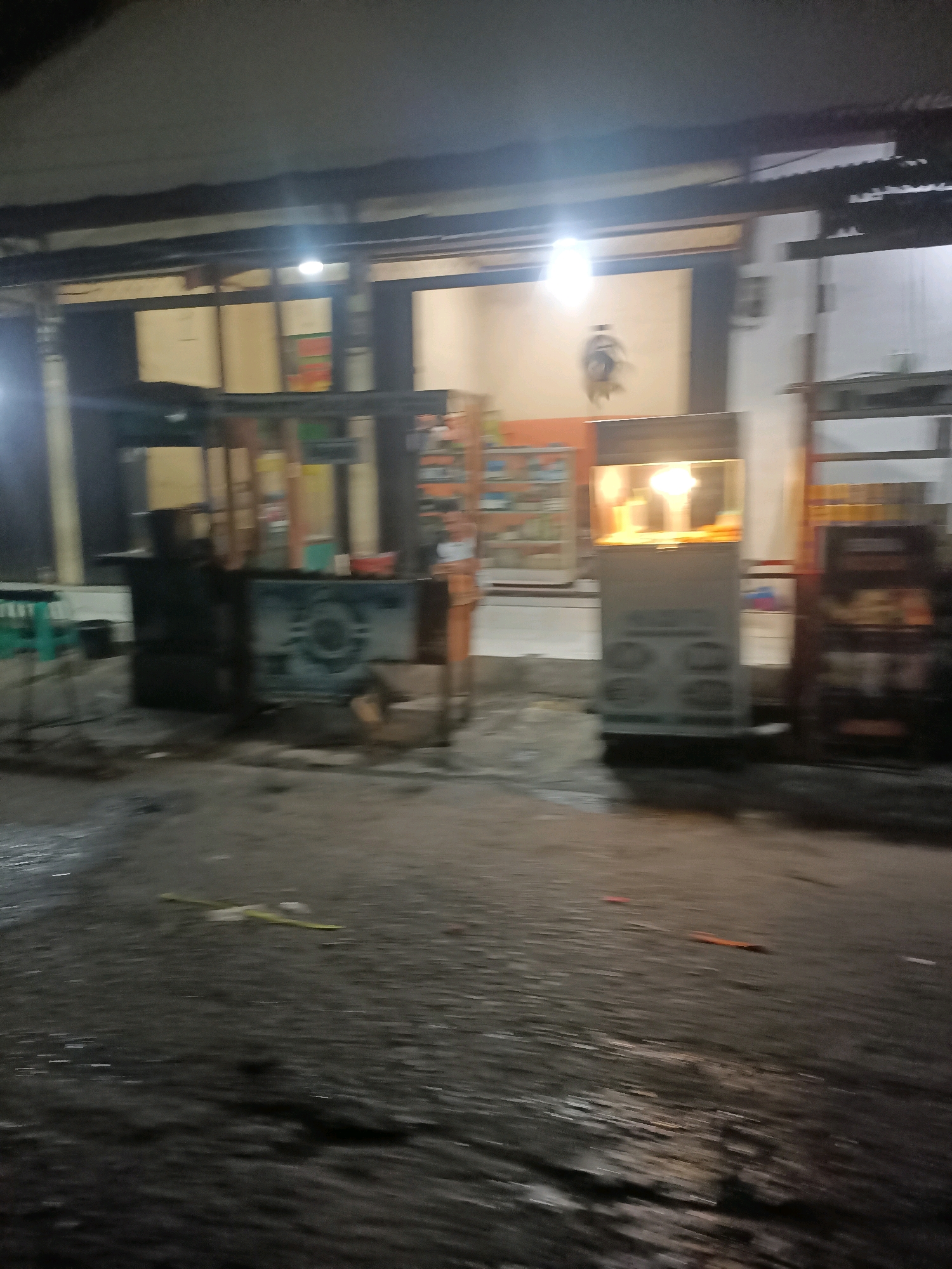 The image depicts a street scene at night in Telagaasih, Indonesia, likely a commercial area. The foreground is a wet, asphalt road reflecting the dim lighting. In the middle ground, there are several food stalls or small shops. One stall on the right has a lit display case, possibly containing food items. To its left, another stall has a dark structure and a visible display or machine with circular patterns. Further to the left, a third stall appears to be a food cart. In the background, there are the facades of buildings, some with open doorways or windows, and awnings or roofs extending over the stalls. The lighting is predominantly from artificial sources, creating a somewhat blurred and atmospheric effect. There are no visible people in the image. The overall impression is that of a typical night market or street food area.