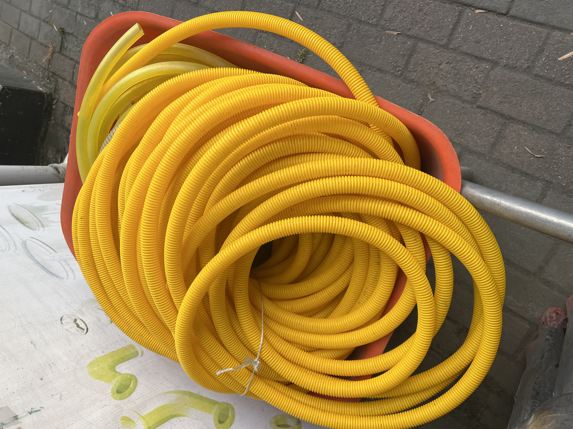 In Batumi, Georgia, a close-up shot captures a bright yellow, corrugated plastic hose coiled tightly within an orange wheelbarrow. The hose, bound by a thin white string, dominates the frame, its ribbed texture creating a visually interesting pattern. To the right of the yellow hose, a smaller, lighter yellow transparent tube is visible, also coiled. The wheelbarrow rests on a dark gray paved surface, possibly a sidewalk or street, with some scattered dry leaves. In the upper left corner, a textured white wall with faint green graffiti and circular blue markings serves as a backdrop. The overall scene appears to be outdoors, likely in a commercial or industrial area given the presence of construction materials like the hose and wheelbarrow.